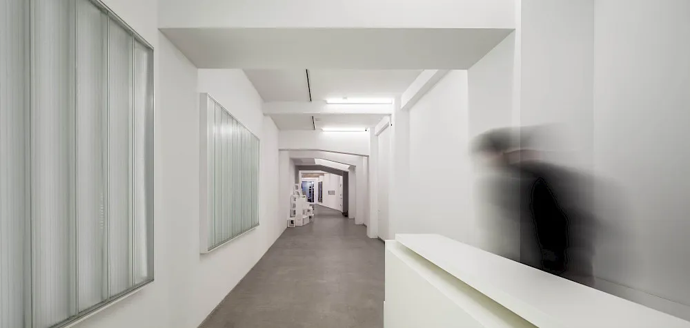 Long white corridor inside a gallery with large framed artworks on the left wall and a blurred figure walking on the right