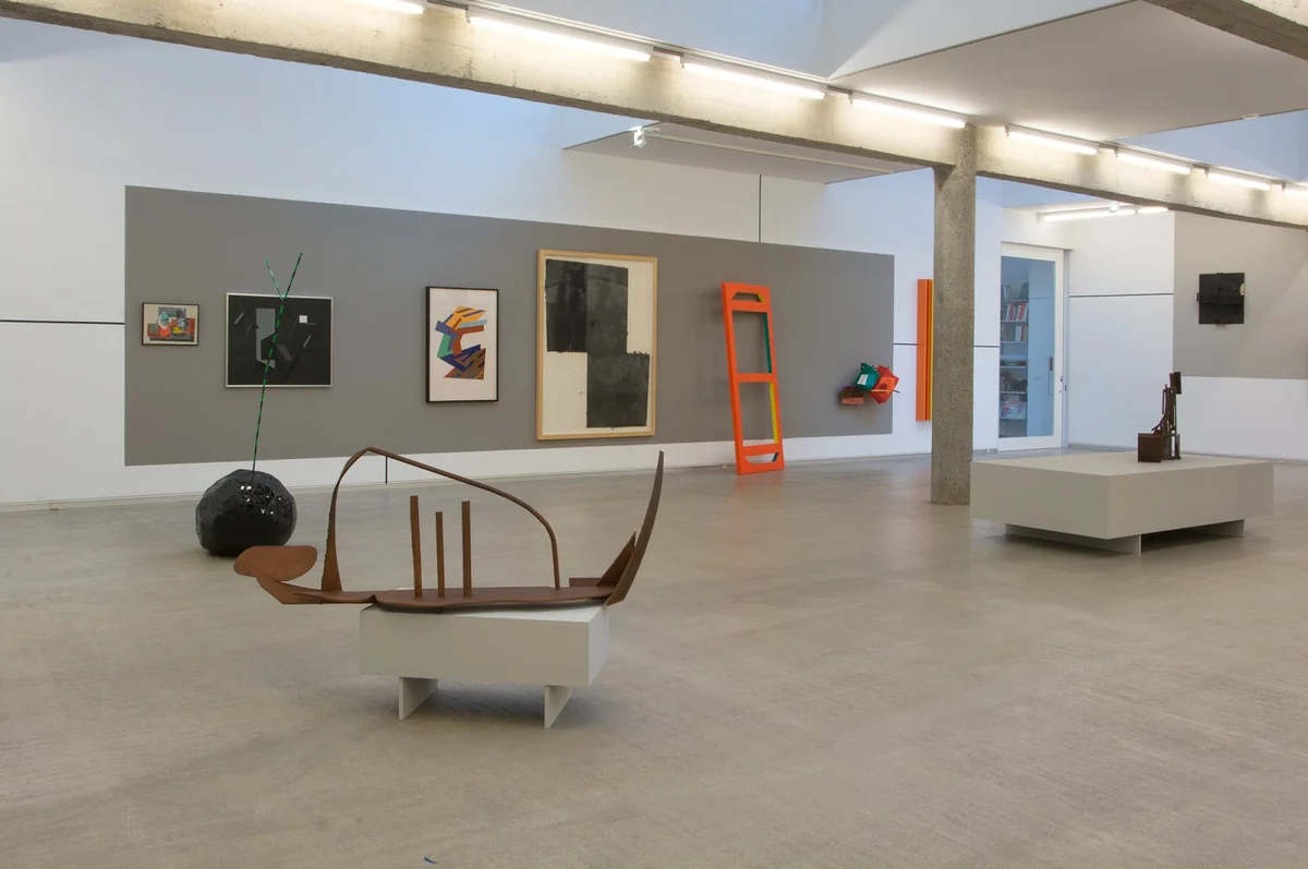 Gallery interior with paintings hung on gray walls and sculptures displayed on white pedestals, including an orange ladder sculpture and a dark spherical object on the floor