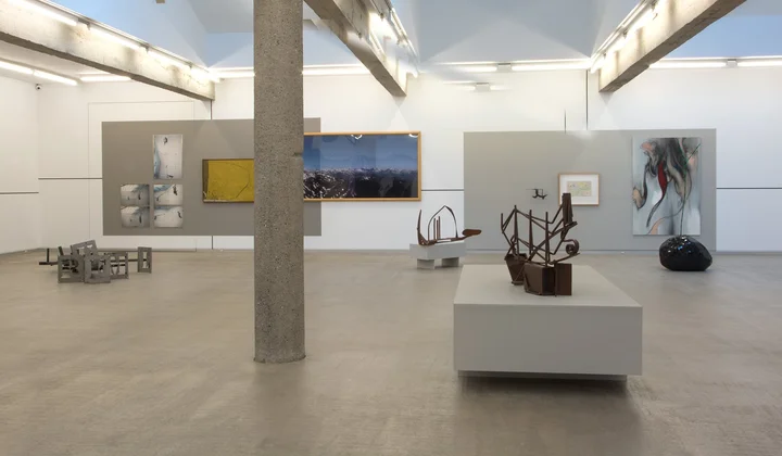 Wide interior view of a contemporary art gallery featuring white walls, a central concrete column, and various paintings and sculptures on display.