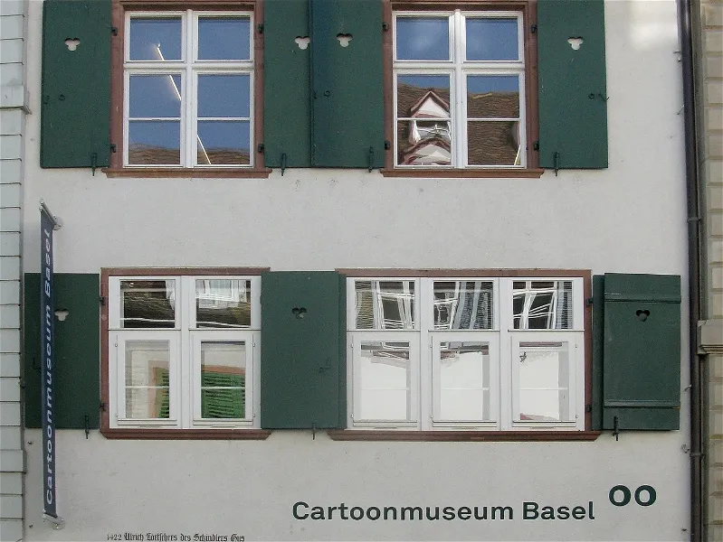 Fachada blanca con contraventanas y ventanas verdes, que muestra el letrero 'Cartoonmuseum Basel' en la parte inferior.