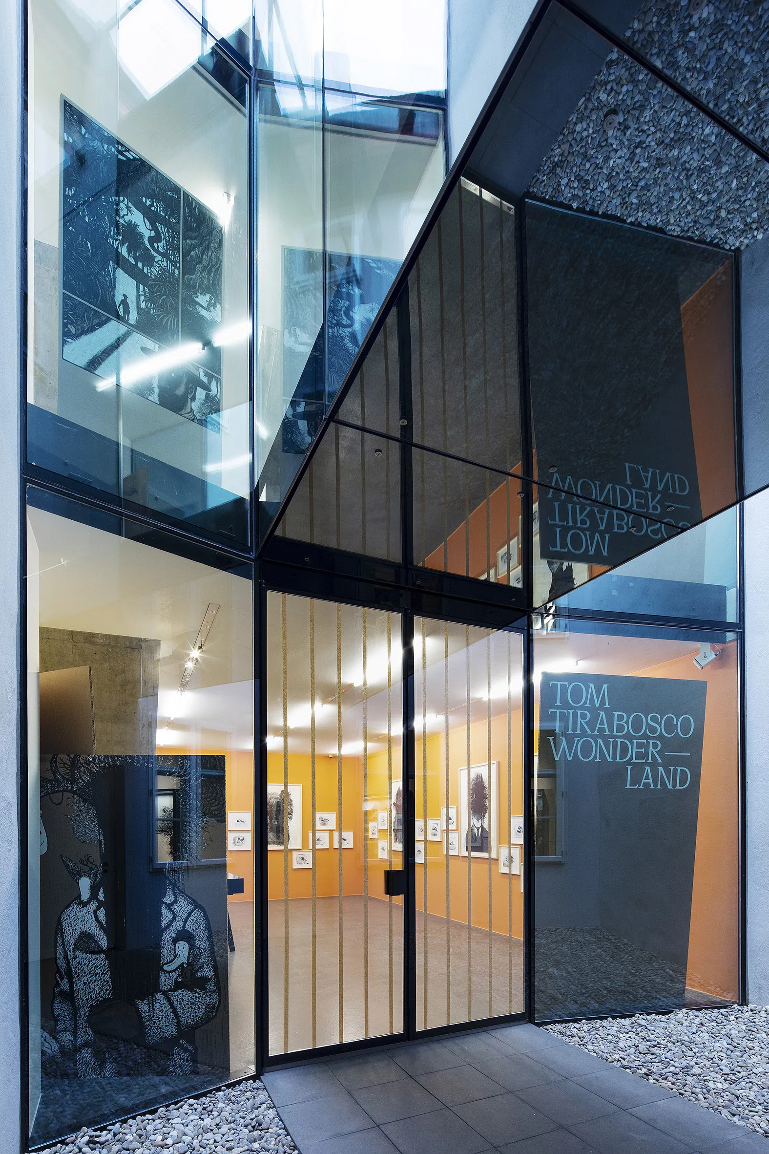Entrada de fachada de cristal del Cartoonmuseum Basel con muros cortina de marco negro, que muestra el texto de la exposición Wonderland de Tom Tirabosco y la galería interior con paredes naranjas y obras de cómic visibles a través del cristal.