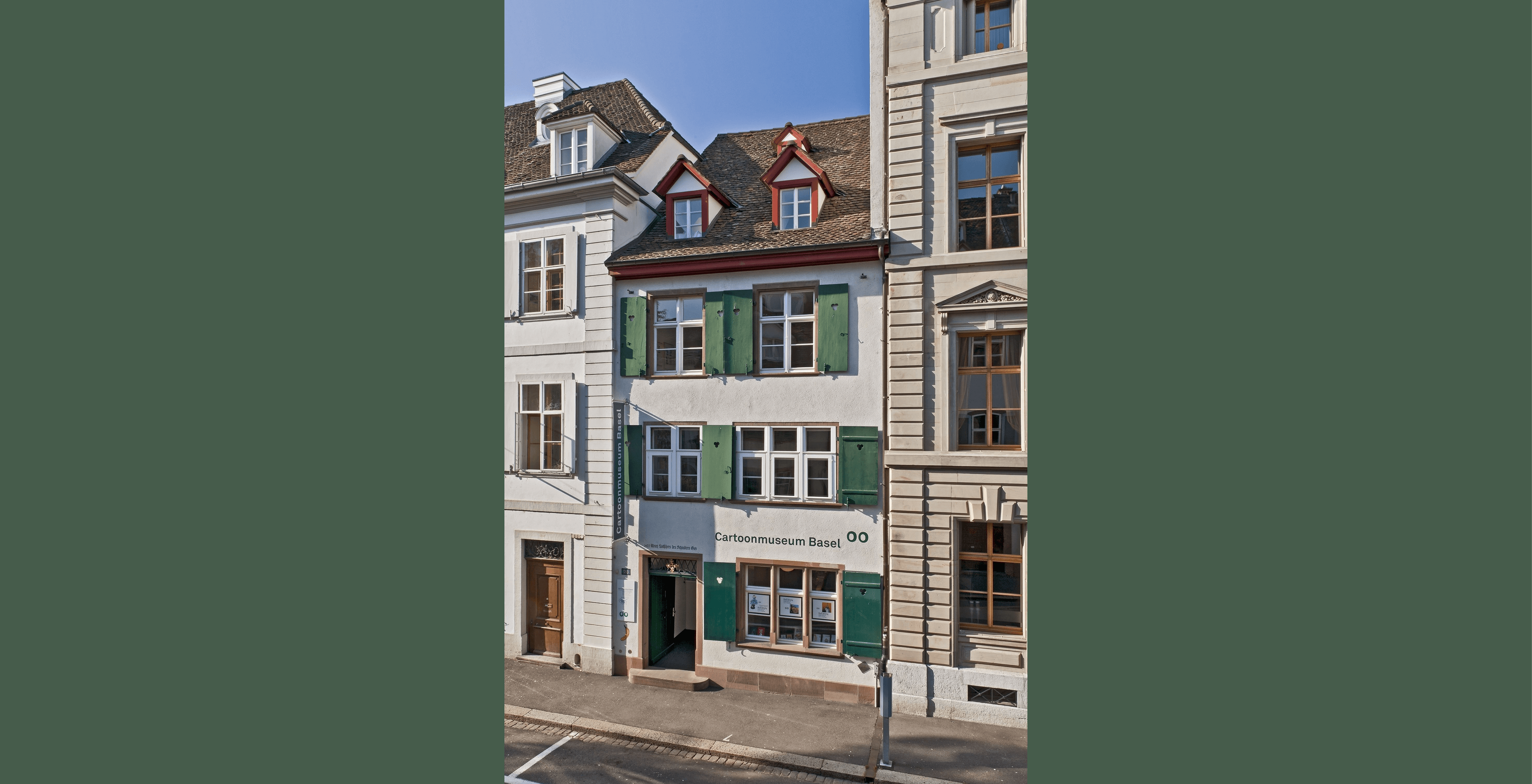 Vista de la fachada del edificio del Cartoonmuseum Basel con contraventanas verdes, paredes blancas y señalización de entrada en una estrecha calle europea