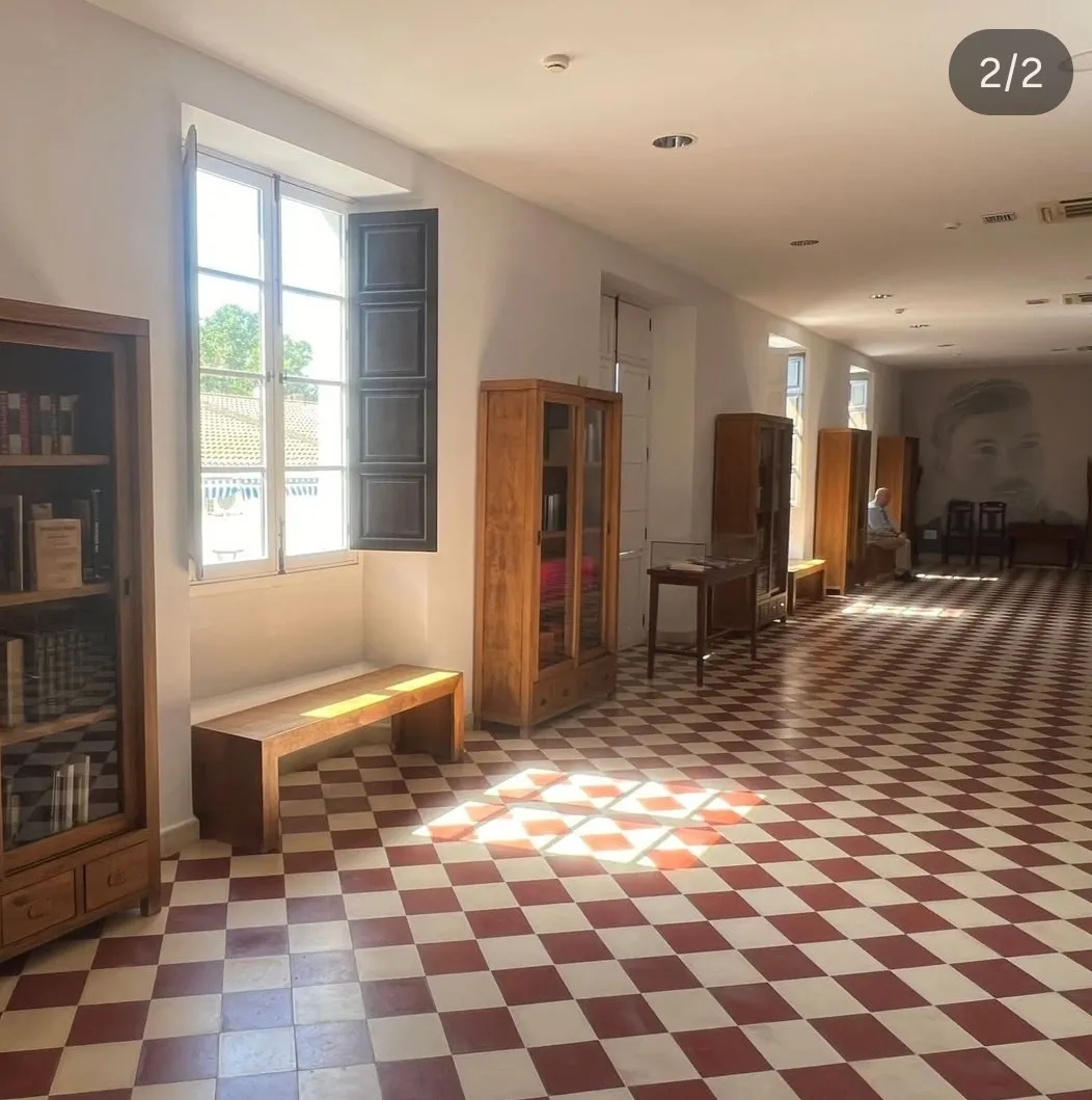Largo vestíbulo interior con suelo de baldosas a cuadros rojos y blancos, vitrinas de madera con puertas de cristal, un banco de madera y ventanas altas con contraventanas oscuras