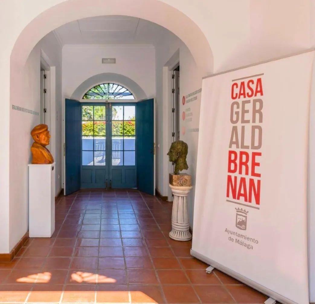 Pasillo interior de Casa Gerald Brenan con suelo de baldosas de terracota, una puerta arqueada blanca, puertas dobles azules que se abren a un jardín, dos bustos sobre pedestales y una gran pancarta blanca que reza Casa Gerald Brenan