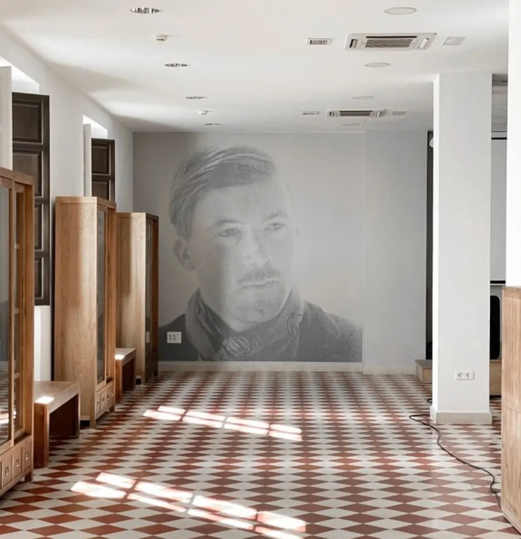 Interior de una sala con suelo de patrón de rombos rojos y blancos, vitrinas de madera a lo largo de la pared izquierda y un gran mural con un retrato en escala de grises en la pared del fondo.