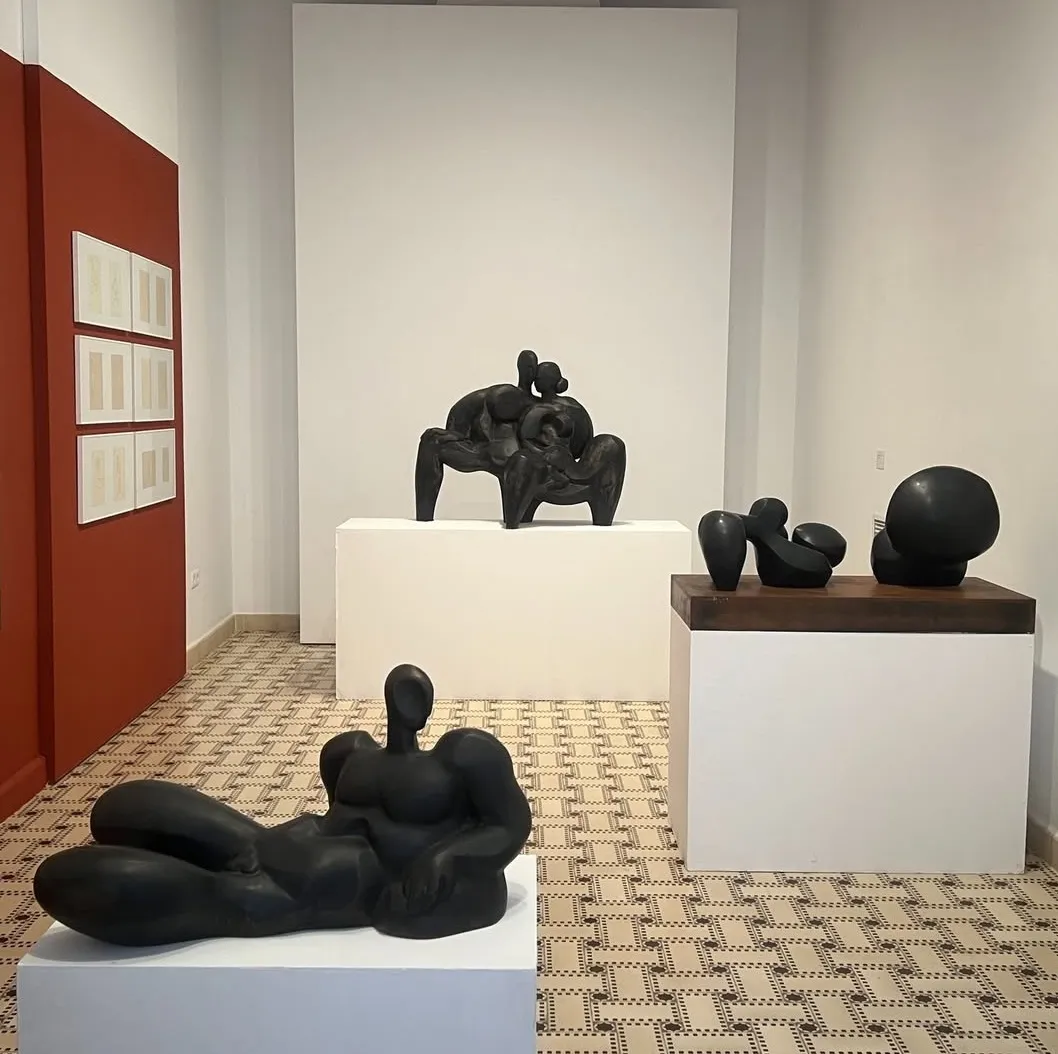 Interior de la galería con esculturas volumétricas negras sobre pedestales blancos, suelo de baldosas con motivos y una pared roja con documentos enmarcados.