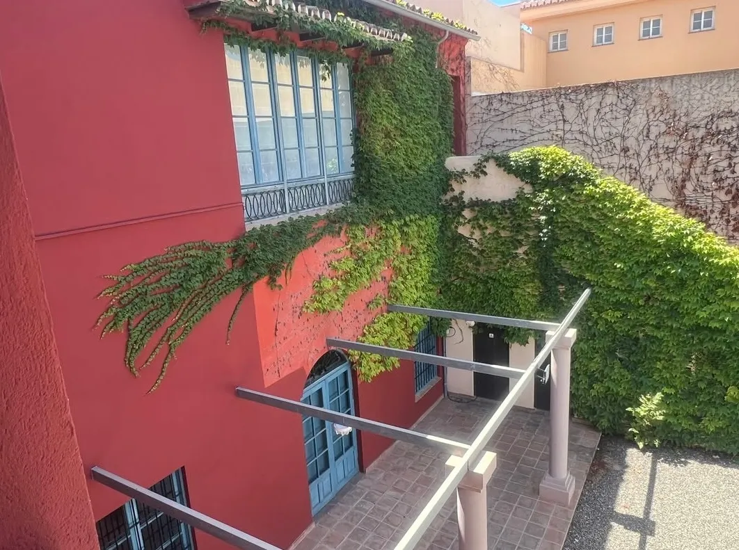 Exterior de edificio rojo cubierto de hiedra verde en la fachada, ventanas con marcos azules y rejas de hierro, estructura de pérgola de metal y un muro de piedra en el patio trasero