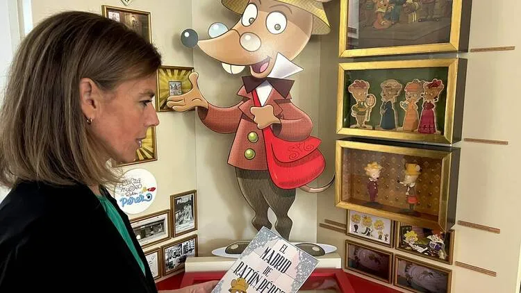 Una mujer mirando una figura grande de un ratón de dibujos animados con un abrigo rojo, rodeada de ilustraciones enmarcadas y un libro.