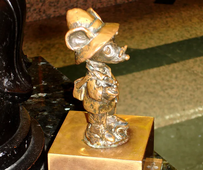 Estatua de bronce de un personaje de ratón con sombrero y mochila sobre una base dorada