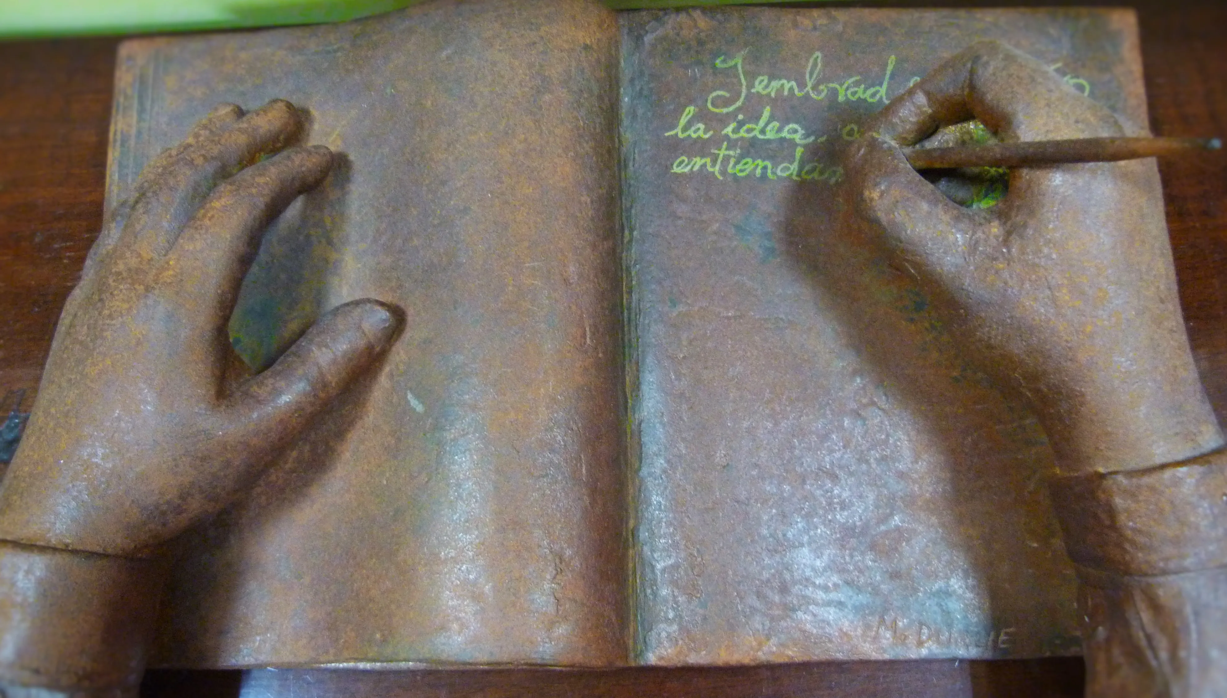 Escultura de bronce de manos escribiendo en un libro abierto con texto en la página