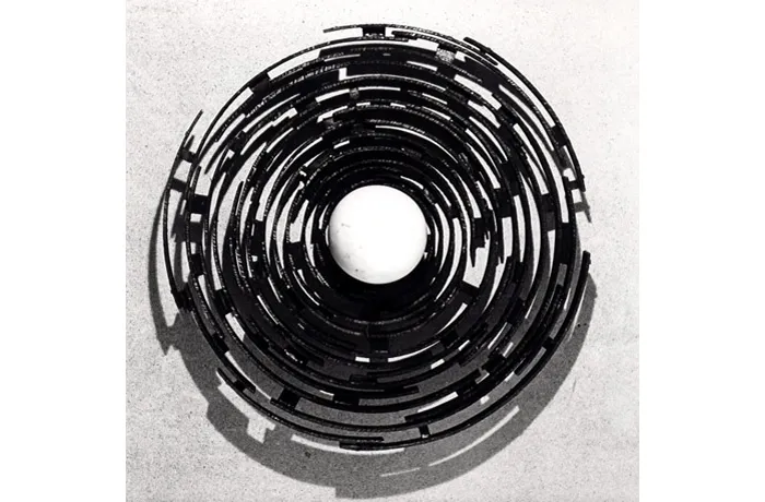 Escultura circular compuesta por segmentos curvos oscuros dispuestos en anillos concéntricos alrededor de un centro blanco, fotografiada en blanco y negro