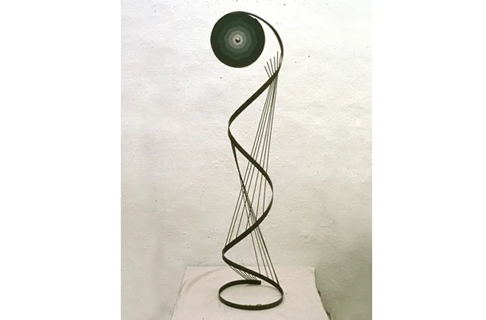 Escultura de metal en espiral con un disco circular oscuro en la parte superior, exhibida sobre un pedestal blanco con un fondo claro