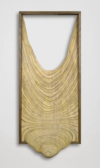 Panel escultórico de madera tallada con líneas curvas concéntricas que forman una profunda forma de U, montado dentro de un marco de madera rectangular sobre una pared blanca.