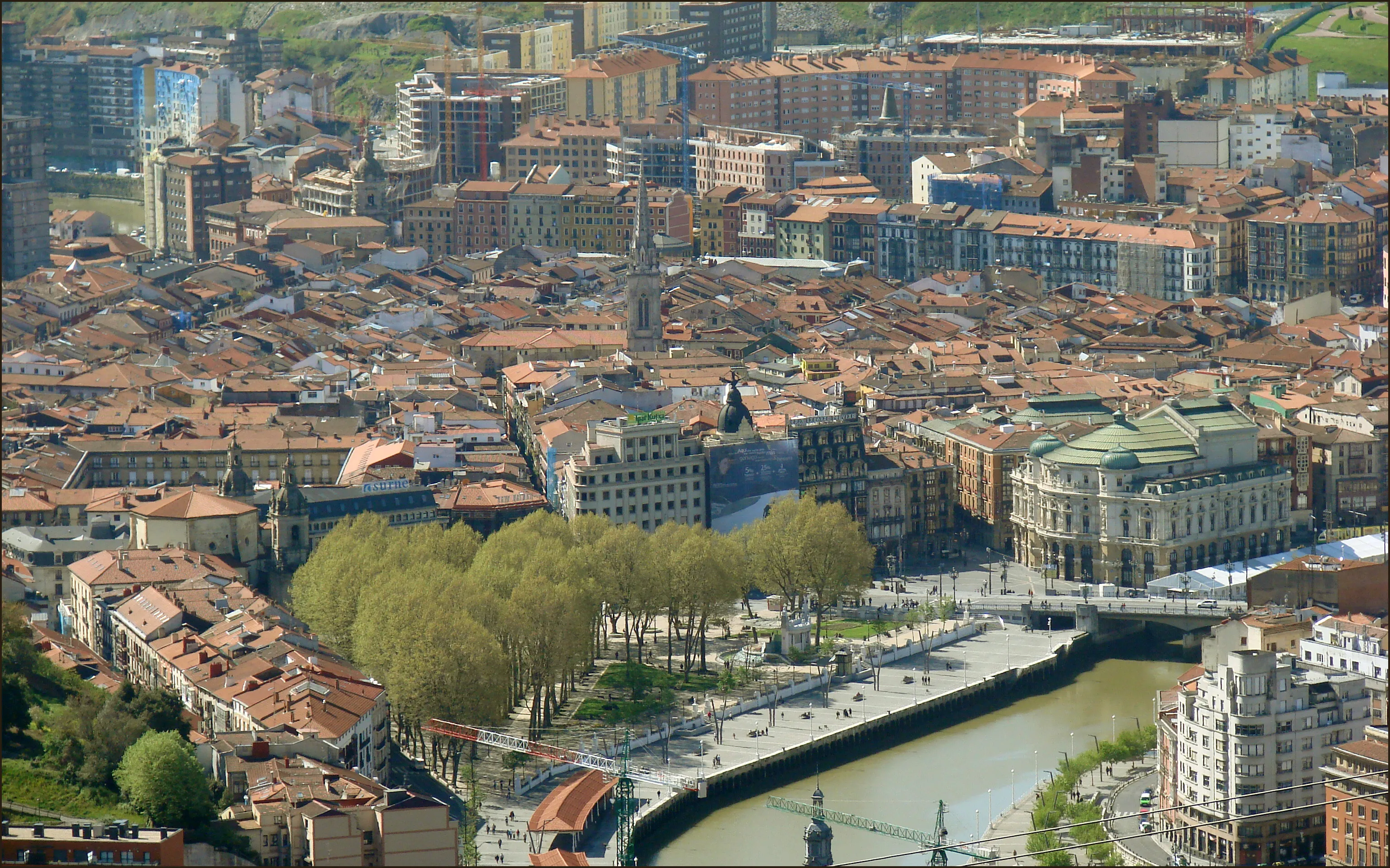 Widok z lotu ptaka na stare miasto w Bilbao, ukazujący gęstą zabudowę z czerwonymi dachami, wieżę kościoła, Teatro Arriaga z zieloną kopułą, wysadzany drzewami bulwar nad rzeką oraz rzekę Nervión wijącą się przez miasto.