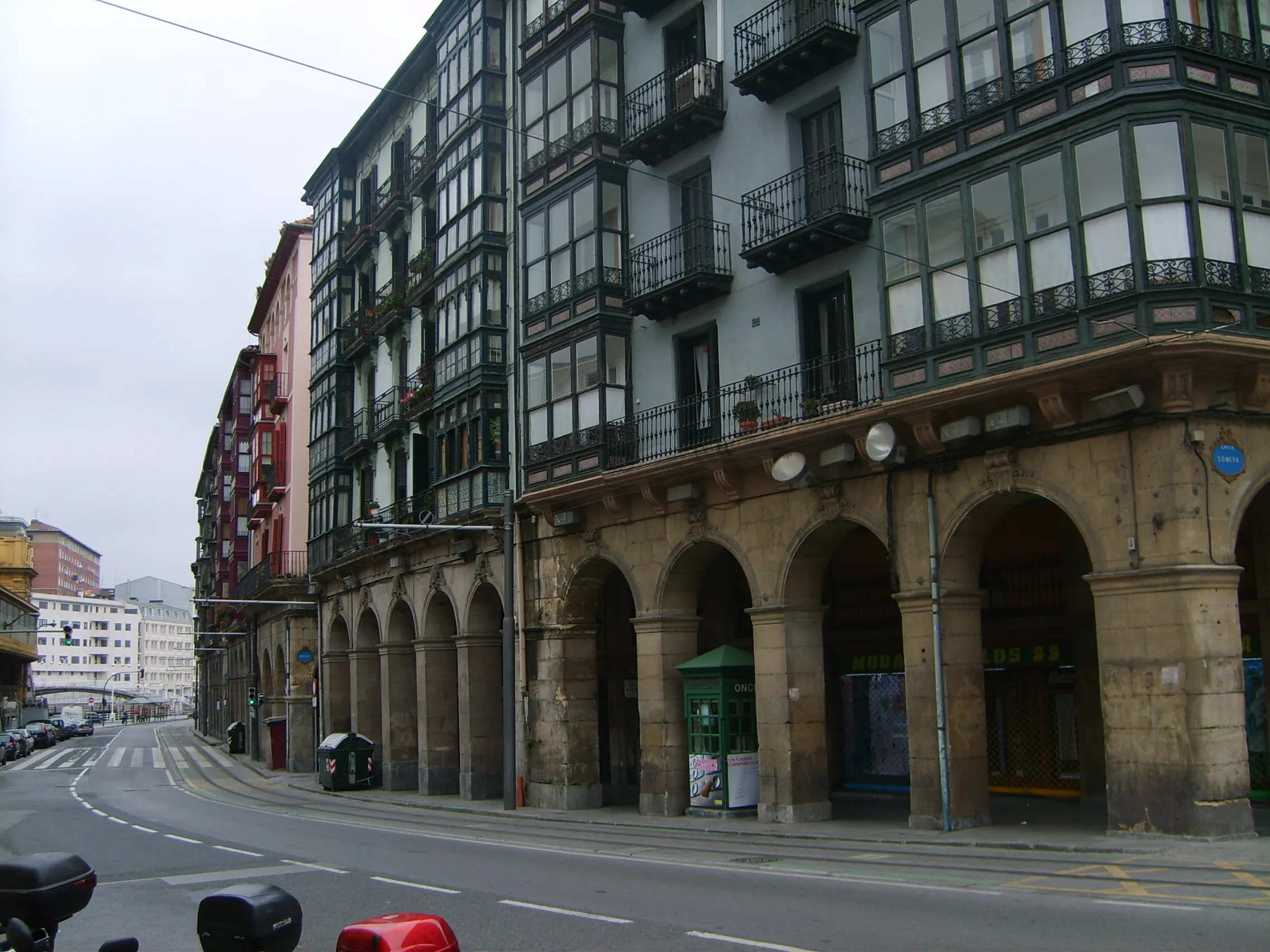 Widok na ulicę La Ribera w dzielnicy Casco Viejo w Bilbao, przedstawiający zabytkowy budynek z kamiennymi łukami na parterze i tradycyjnymi drewnianymi balkonami w stylu baskijskim na wyższych piętrach.