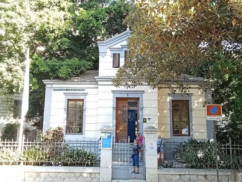 Pequeño pabellón de color blanco y amarillo con puerta de entrada azul y verja de hierro, rodeado de árboles, con visitantes parados en la puerta