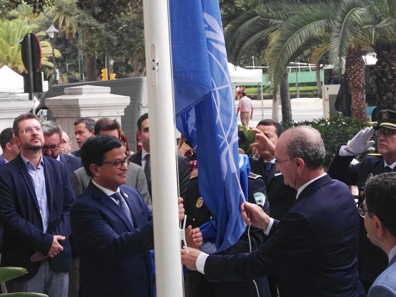 Grupo de personas izando una bandera azul de las Naciones Unidas en un asta blanca al aire libre con palmeras de fondo