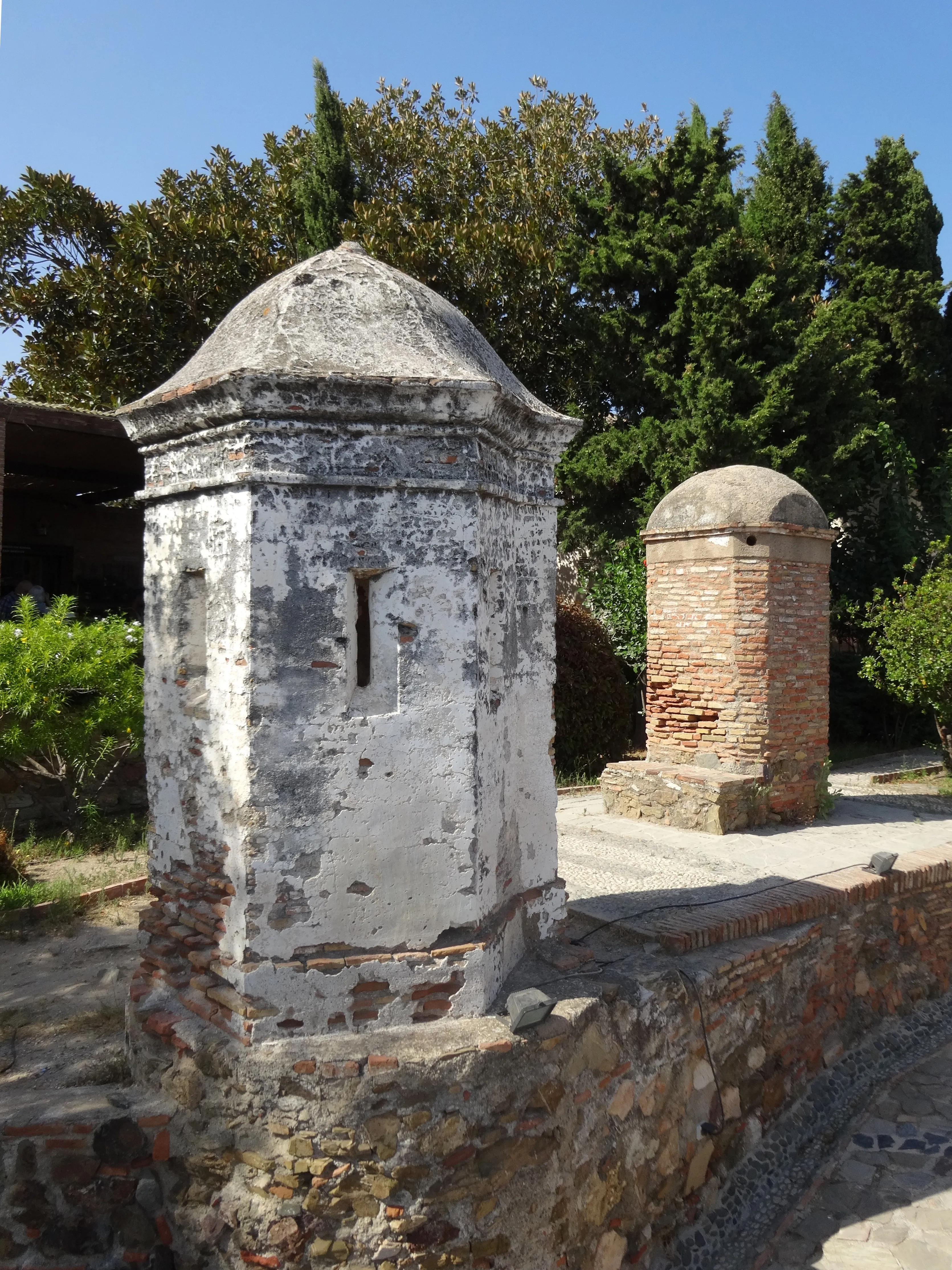 Dos pequeñas garitas con cúpula sobre una muralla de piedra, una encalada con una estrecha ventana y la otra de ladrillo visto, con árboles verdes al fondo bajo un cielo azul.