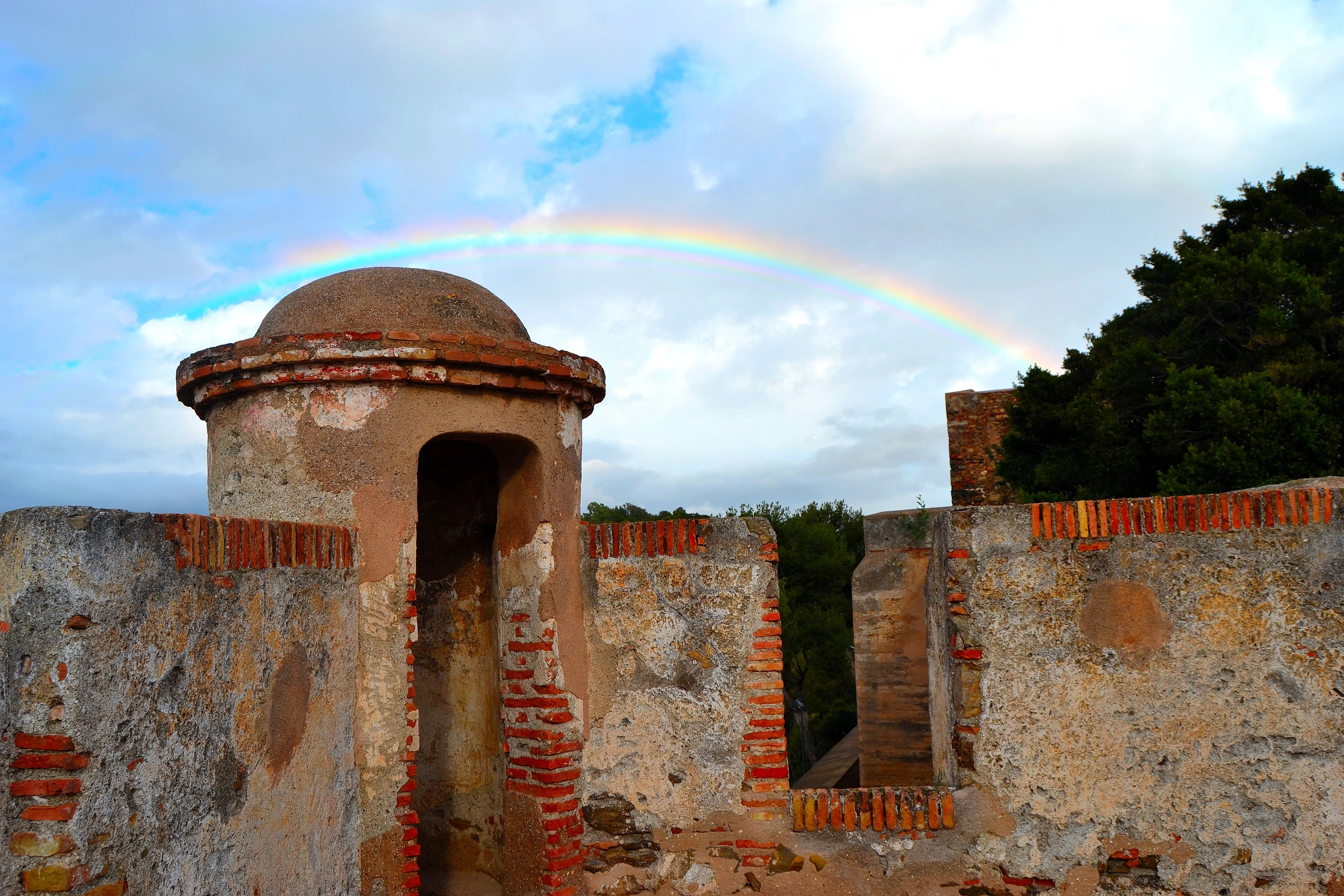 Muros de fortaleza de piedra con una torre abovedada y detalles de ladrillo visto, con un arcoíris arqueándose sobre un cielo azul nublado de fondo.