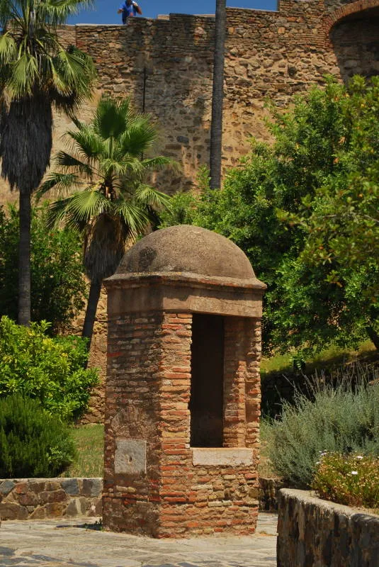 Una pequeña garita de ladrillo con techo abovedado se alza entre palmeras y vegetación, con los muros de piedra de la fortaleza elevándose al fondo en el Castillo de Gibralfaro.