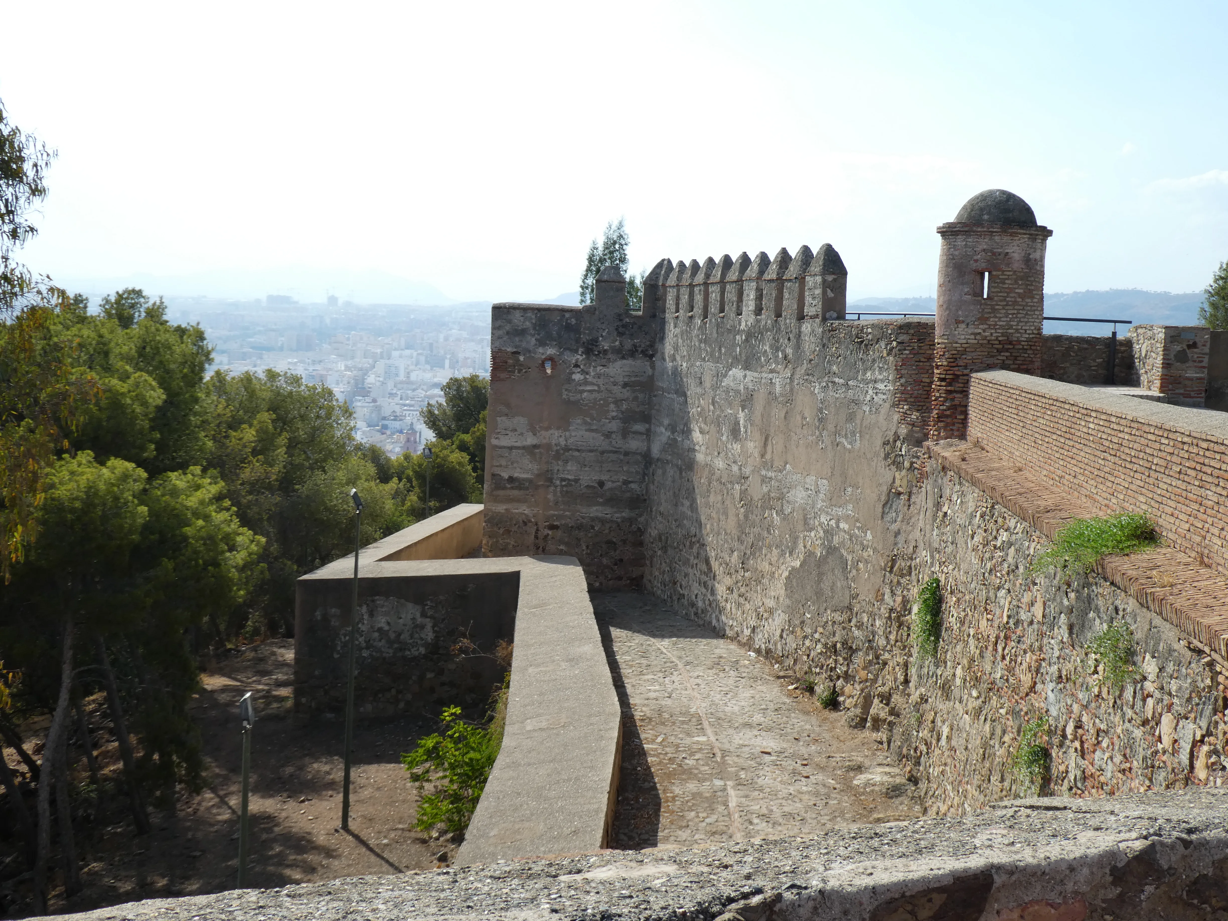 Murallas de piedra y un paseo a lo largo de las murallas del Castillo de Gibralfaro, con vistas a la ciudad y al mar de fondo.