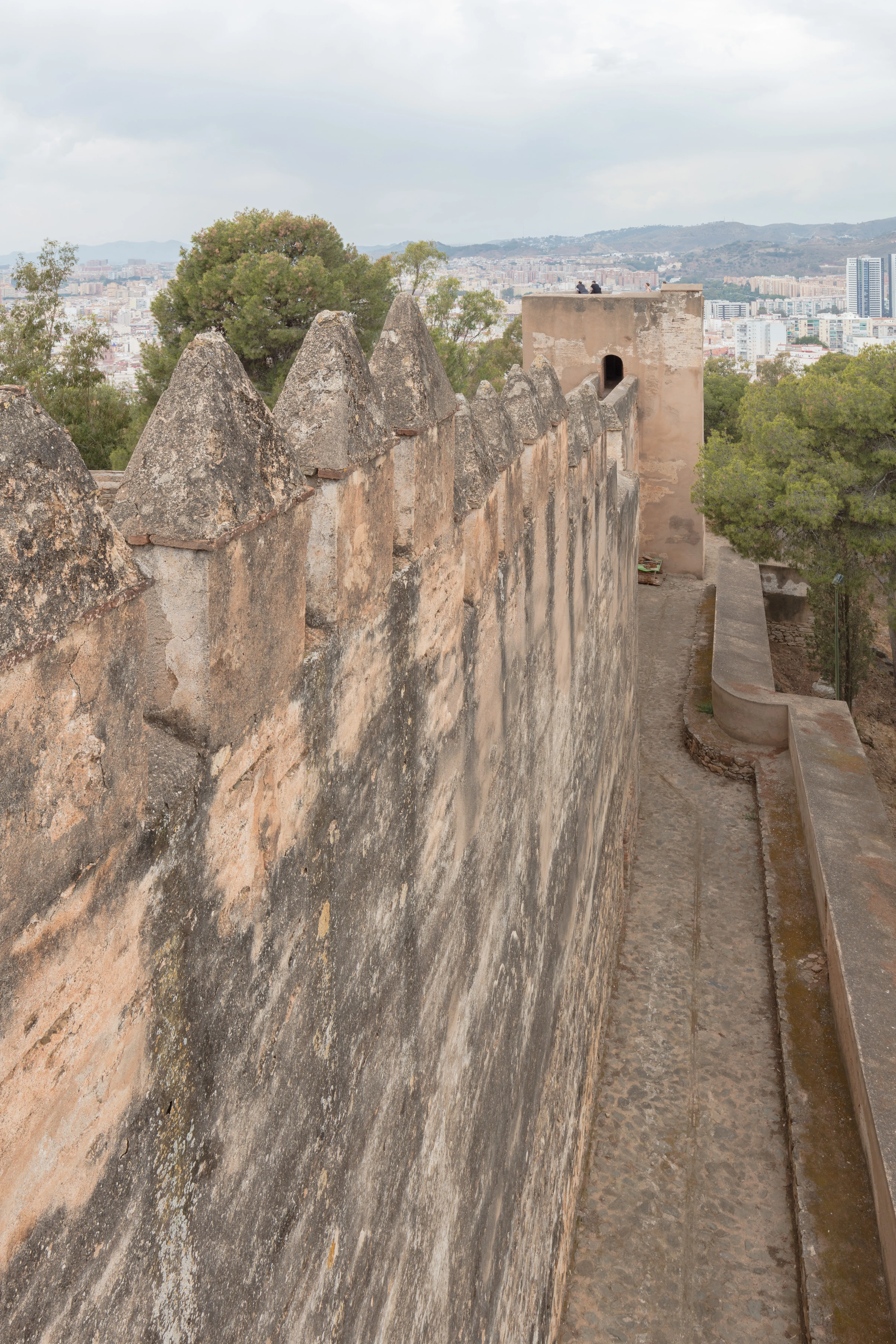 Murallas de piedra con almenas cónicas a lo largo de los muros del castillo, con el paisaje urbano de Málaga y las colinas visibles al fondo