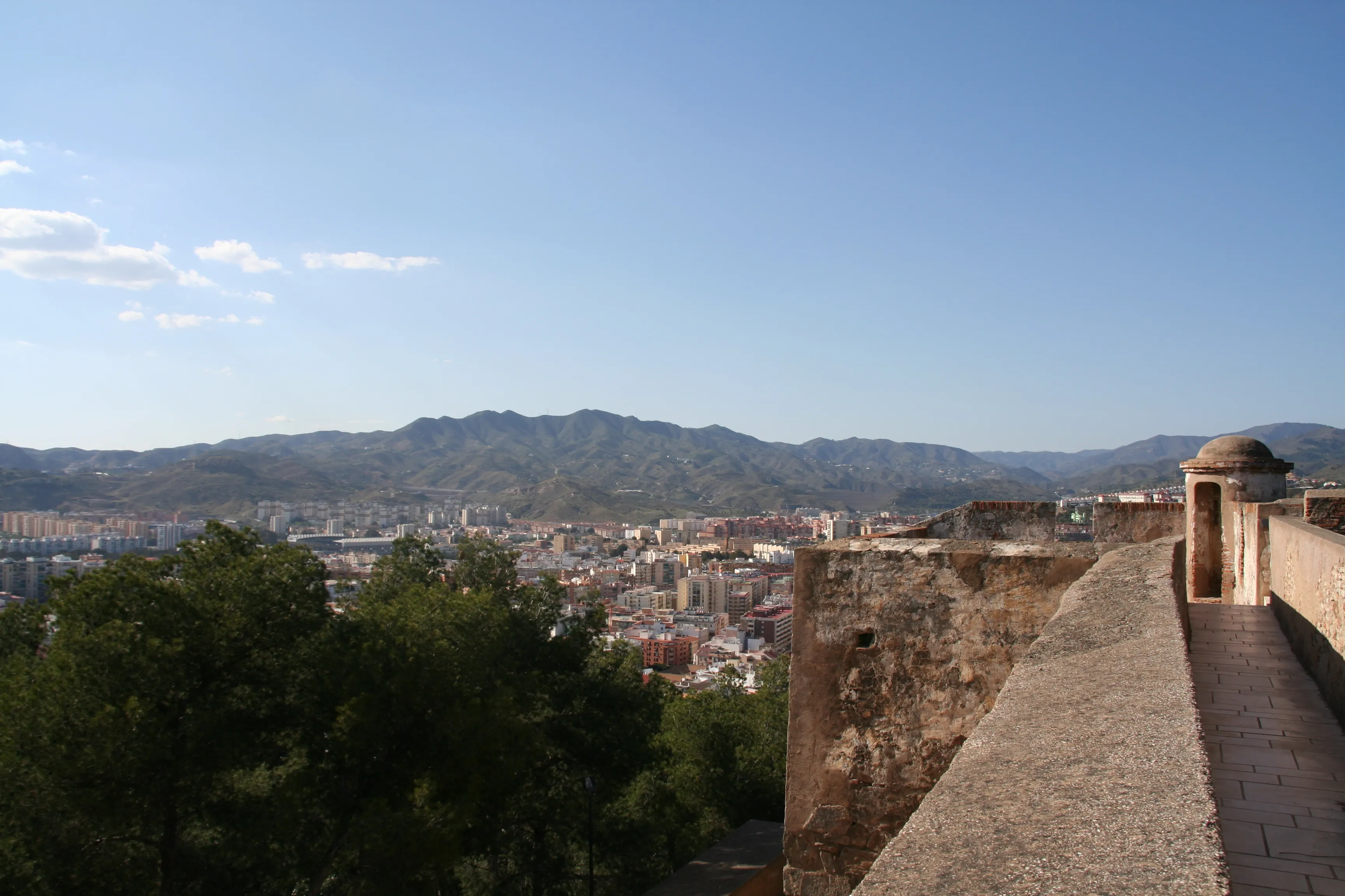Muralla de piedra del Castillo de Gibralfaro con una pequeña torre abovedada, con vistas a la ciudad de Málaga y a las montañas bajo un cielo azul