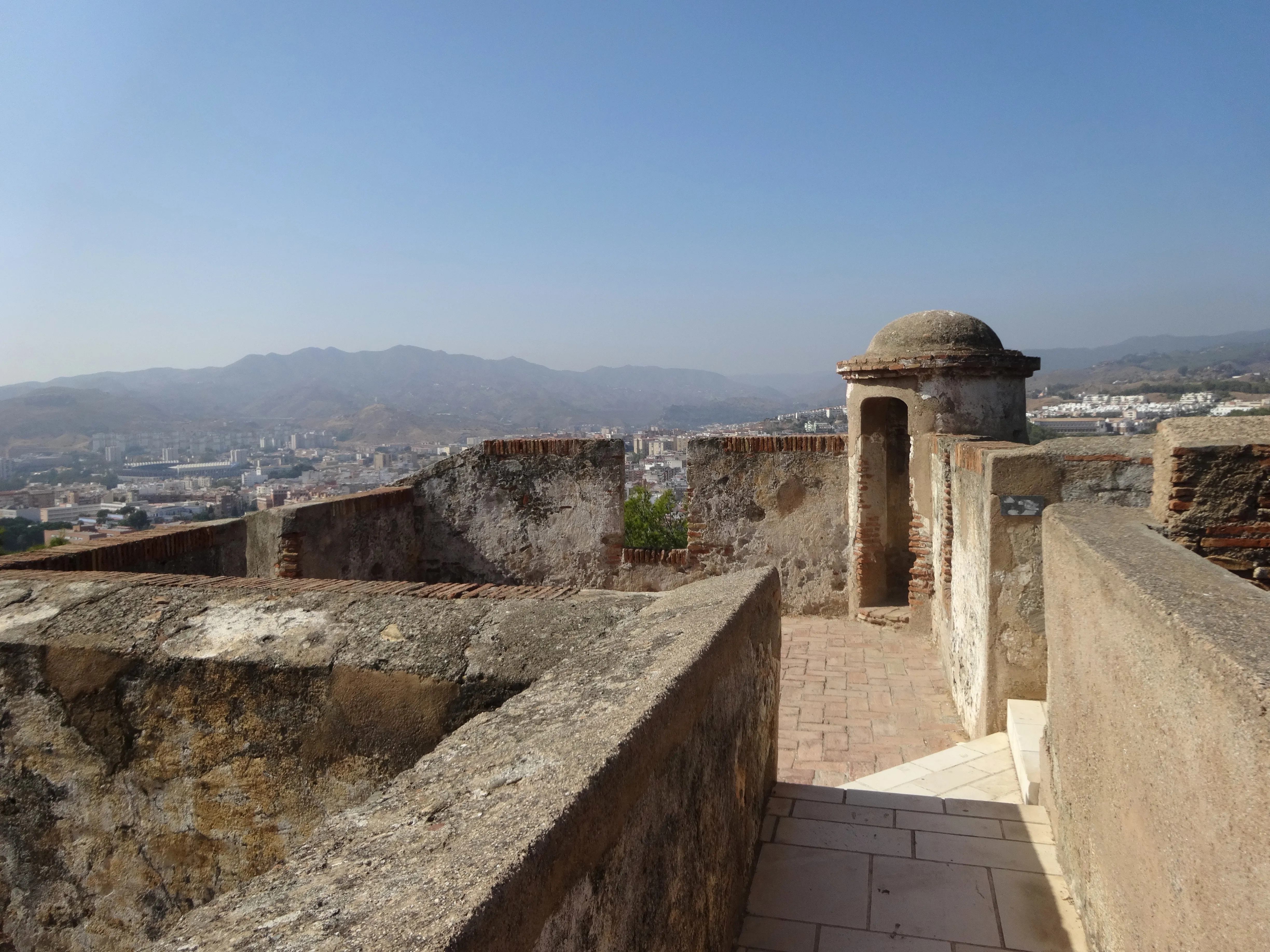 Murallas de piedra y una garita abovedada en el Castillo de Gibralfaro con vistas a la ciudad de Málaga y sus montañas bajo un cielo azul despejado.