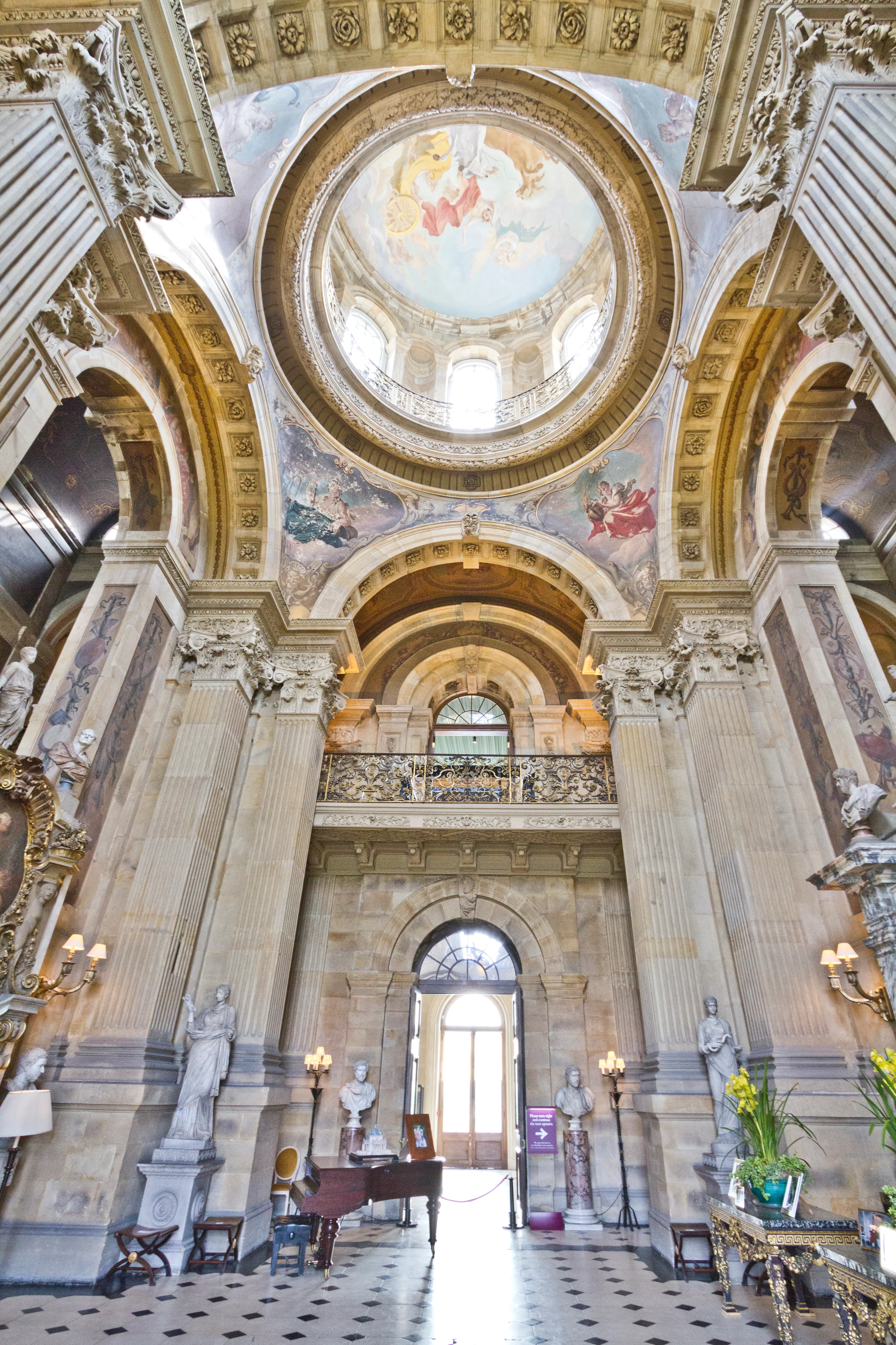 Het interieur van de Great Hall in Castle Howard met een grote beschilderde koepel, hoge klassieke zuilen, marmeren beelden, wandkandelaars en een vleugel op een zwart-witte geblokte vloer.