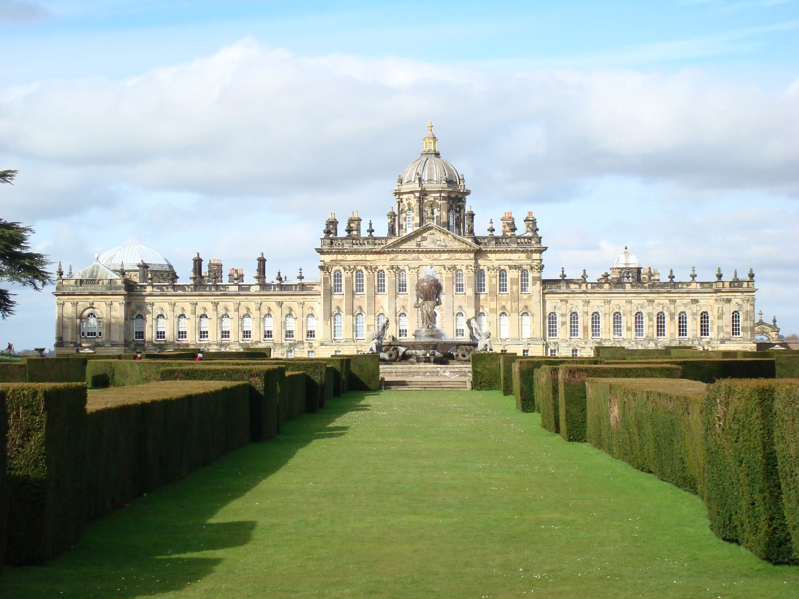 De barokke gevel van Castle Howard met centrale koepel, gezien over een strak gazon geflankeerd door hoge heggen die naar een fontein leiden