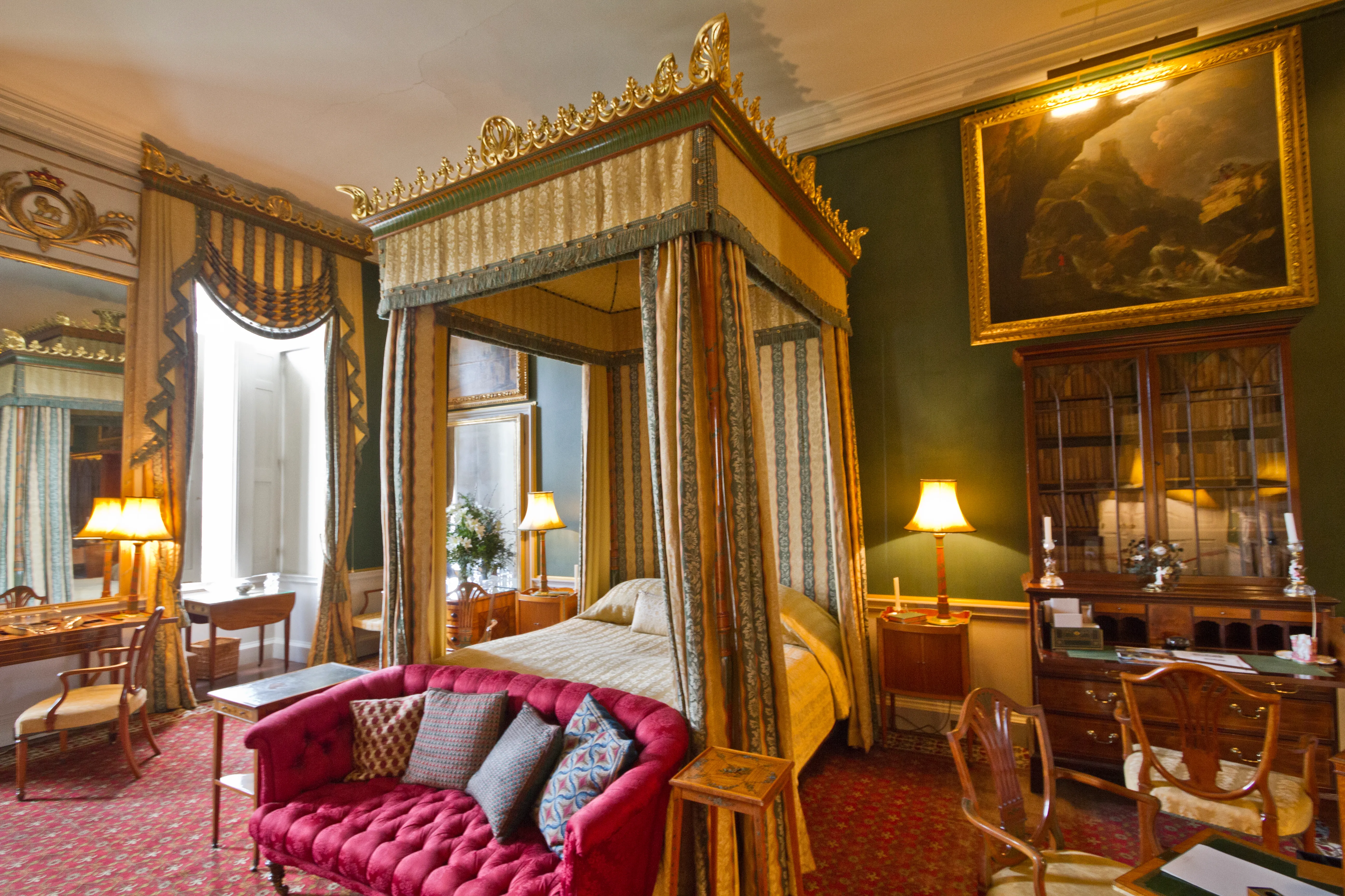Rijkversierd hemelbed met goudkleurige en groene gordijnen in een grandioze slaapkamer met een rode gecapitonneerde bank en een groot landschapsschilderij.