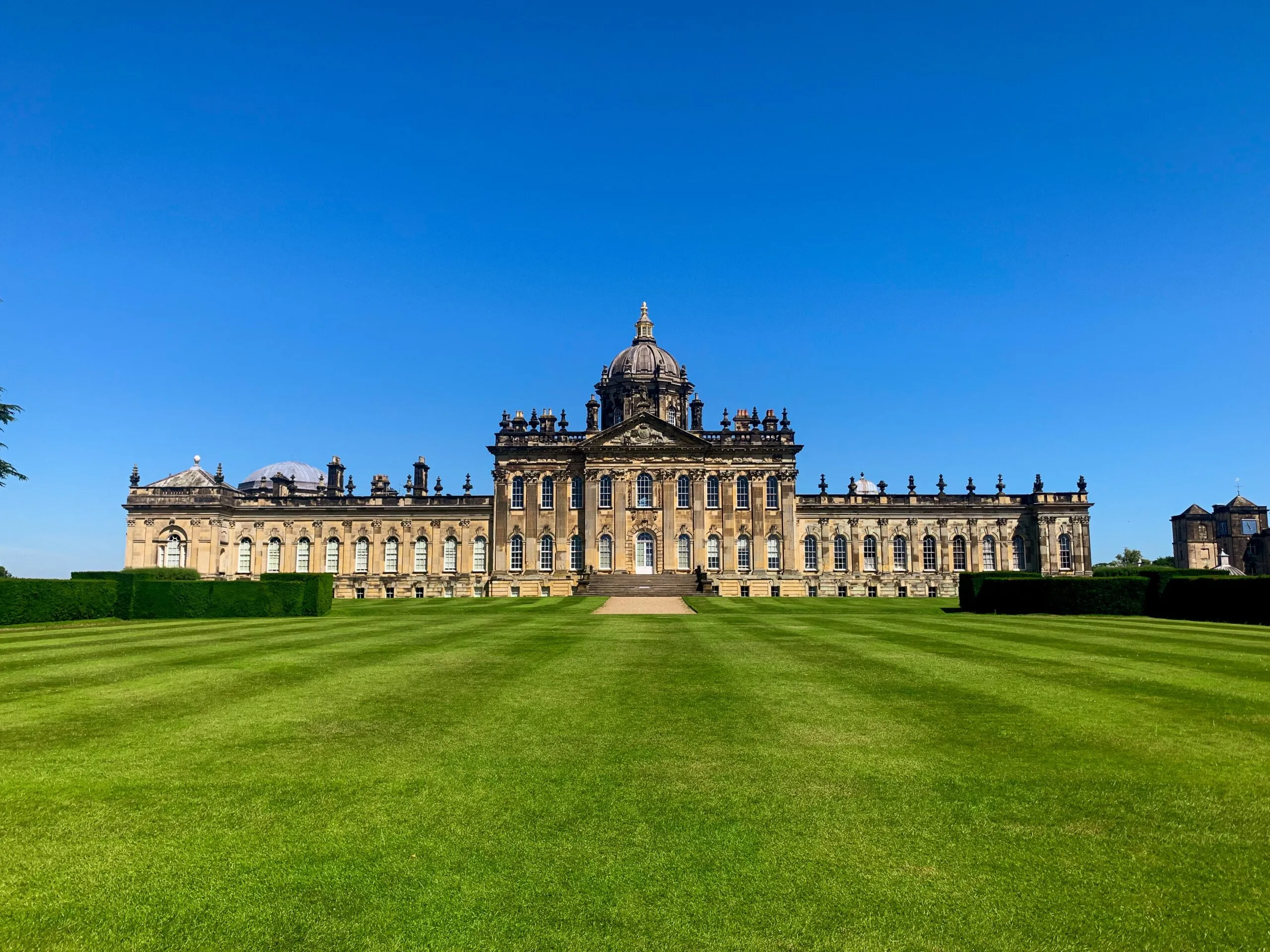De grandioze barokke gevel van Castle Howard met zijn centrale koepel, geflankeerd door symmetrische vleugels, gezien over een breed, verzorgd gazon onder een strakblauwe lucht