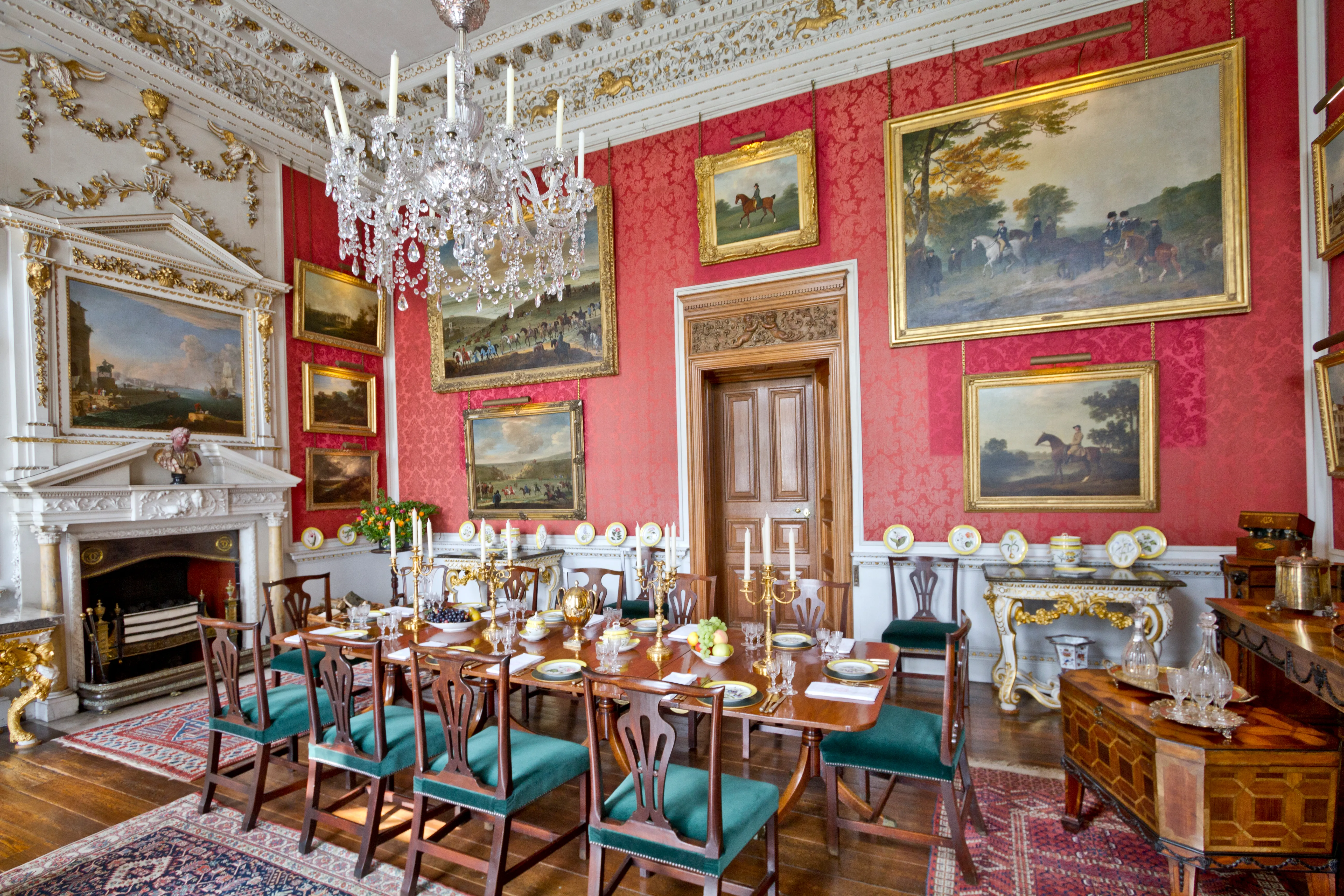 Crimson Dining Room in Castle Howard met rode muren, kristallen kroonluchter, marmeren schouw en een lange eettafel gedekt met stoelen bekleed met blauwgroene stof, omringd door ingelijste landschapsschilderijen.