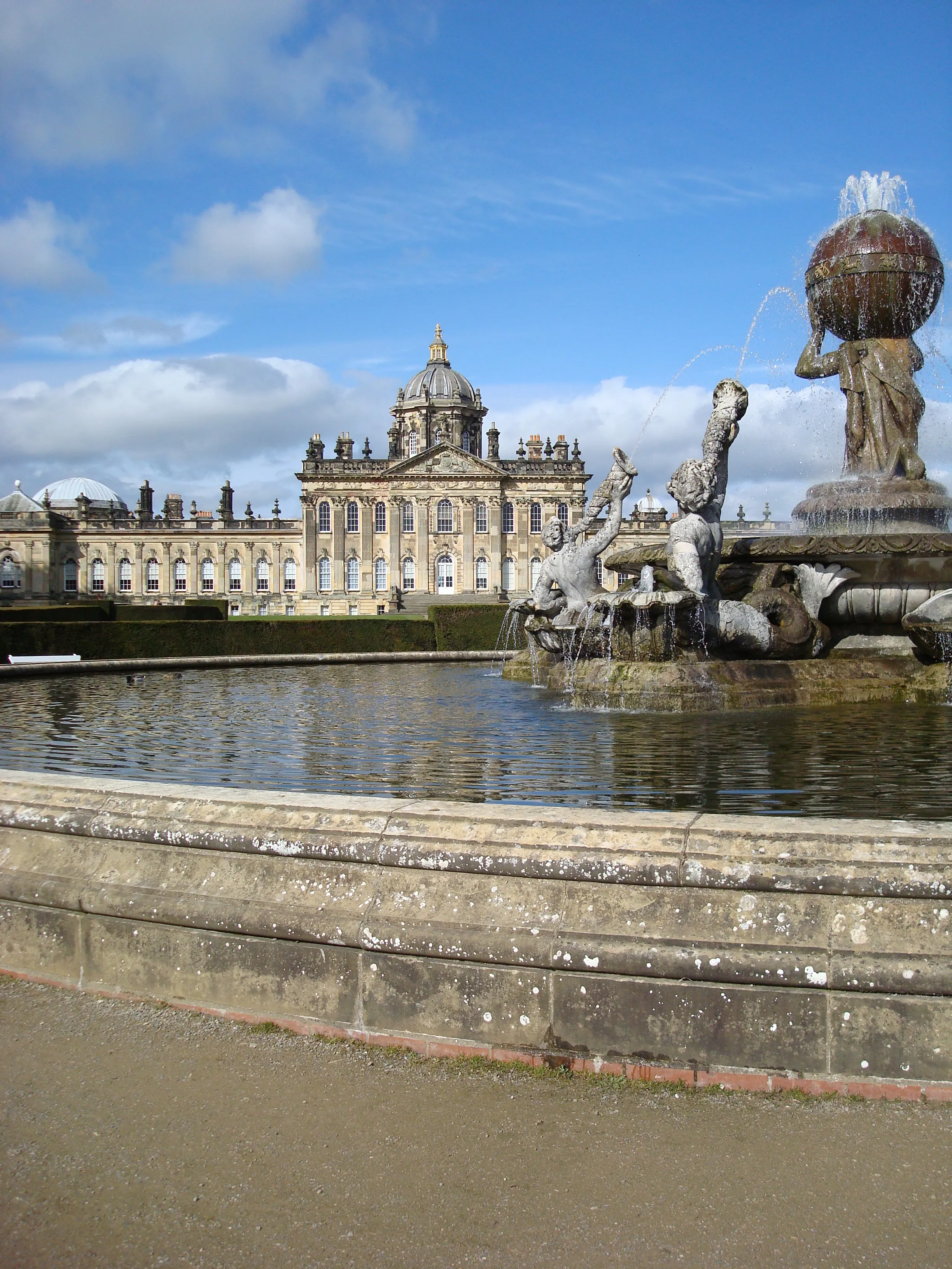 De barokke gevel van Castle Howard met centrale koepel, gezien vanaf de overkant van een spiegelvijver met de sierlijke Atlas Fountain op de voorgrond