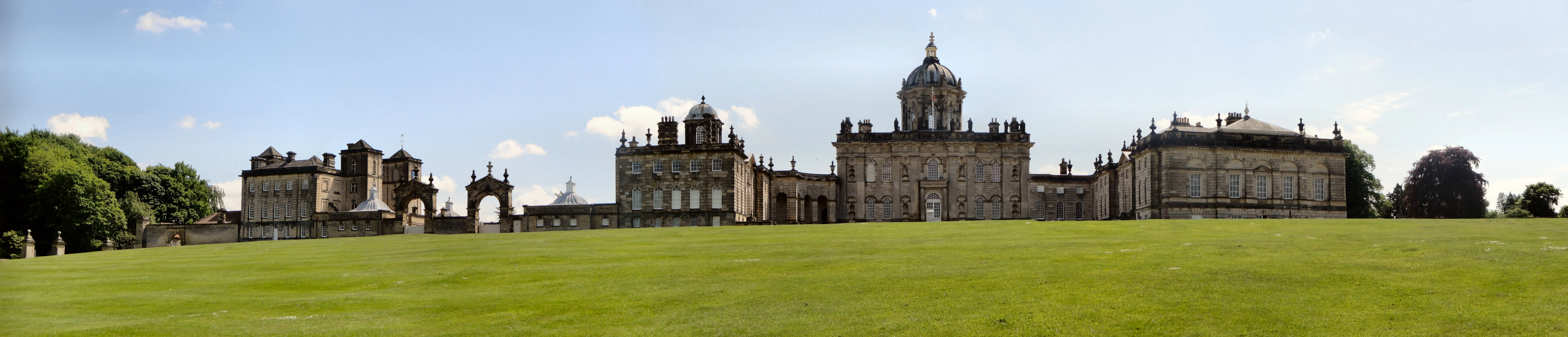Weids panoramisch uitzicht op de barokke gevel van Castle Howard met de centrale koepel, de flankerende vleugels en het uitgestrekte groene gazon onder een blauwe lucht met verspreide wolken.