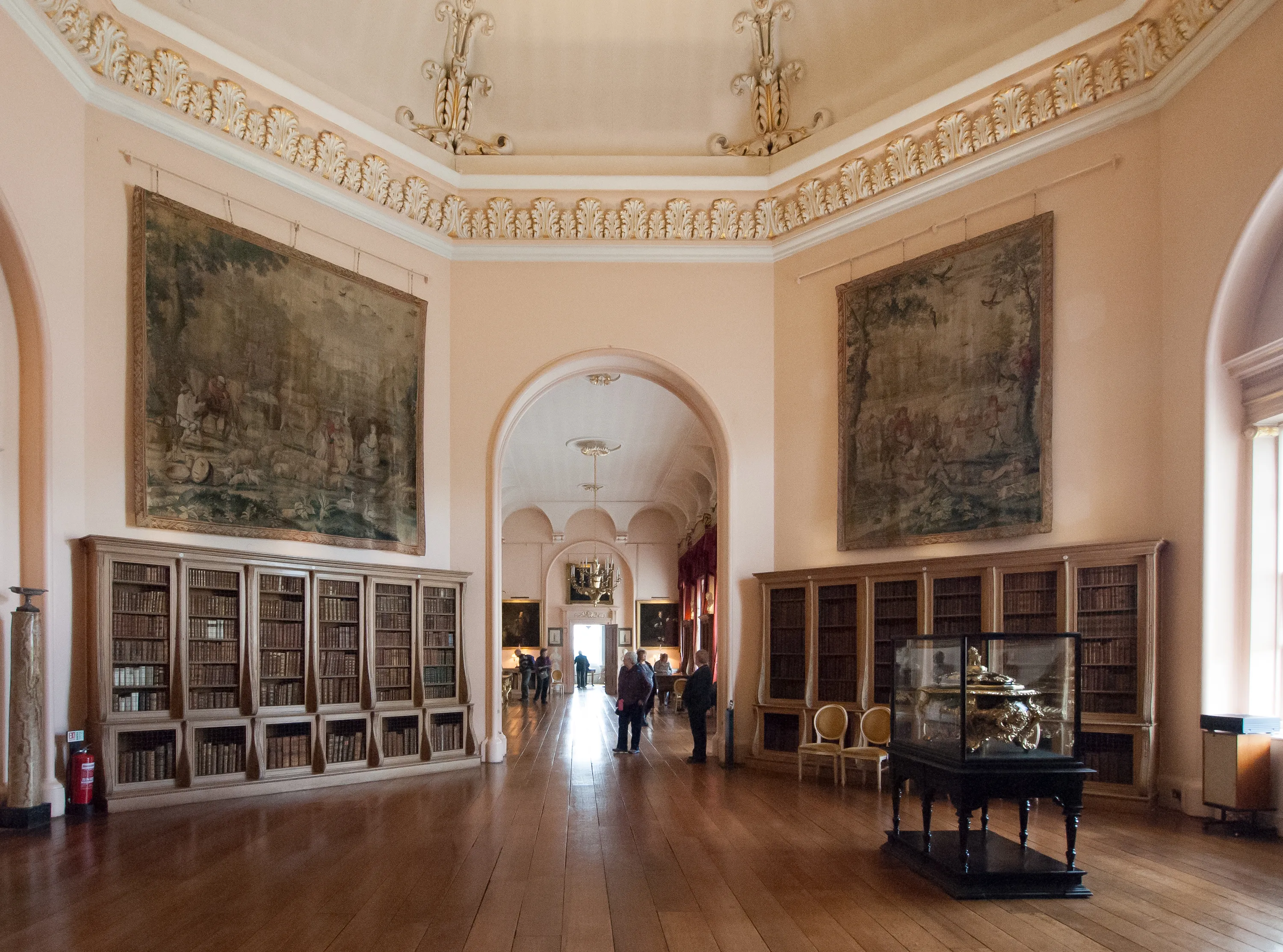 Grote binnenruimte in Castle Howard met roze muren, rijkelijk versierde plafondlijsten, grote wandtapijten, kamerhoge boekenplanken en een boogvormige deuropening die leidt naar een gang met bezoekers.