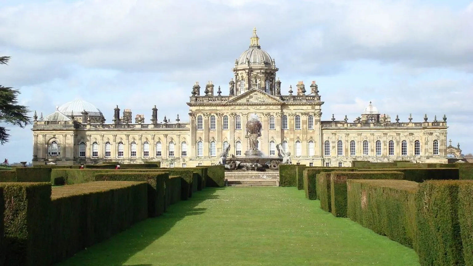 Breed buitenaanzicht van de barokke gevel van Castle Howard met centrale koepel, geflankeerd door symmetrische vleugels en formele omhaagde tuinpaden die naar een centraal standbeeld leiden onder een halfbewolkte hemel