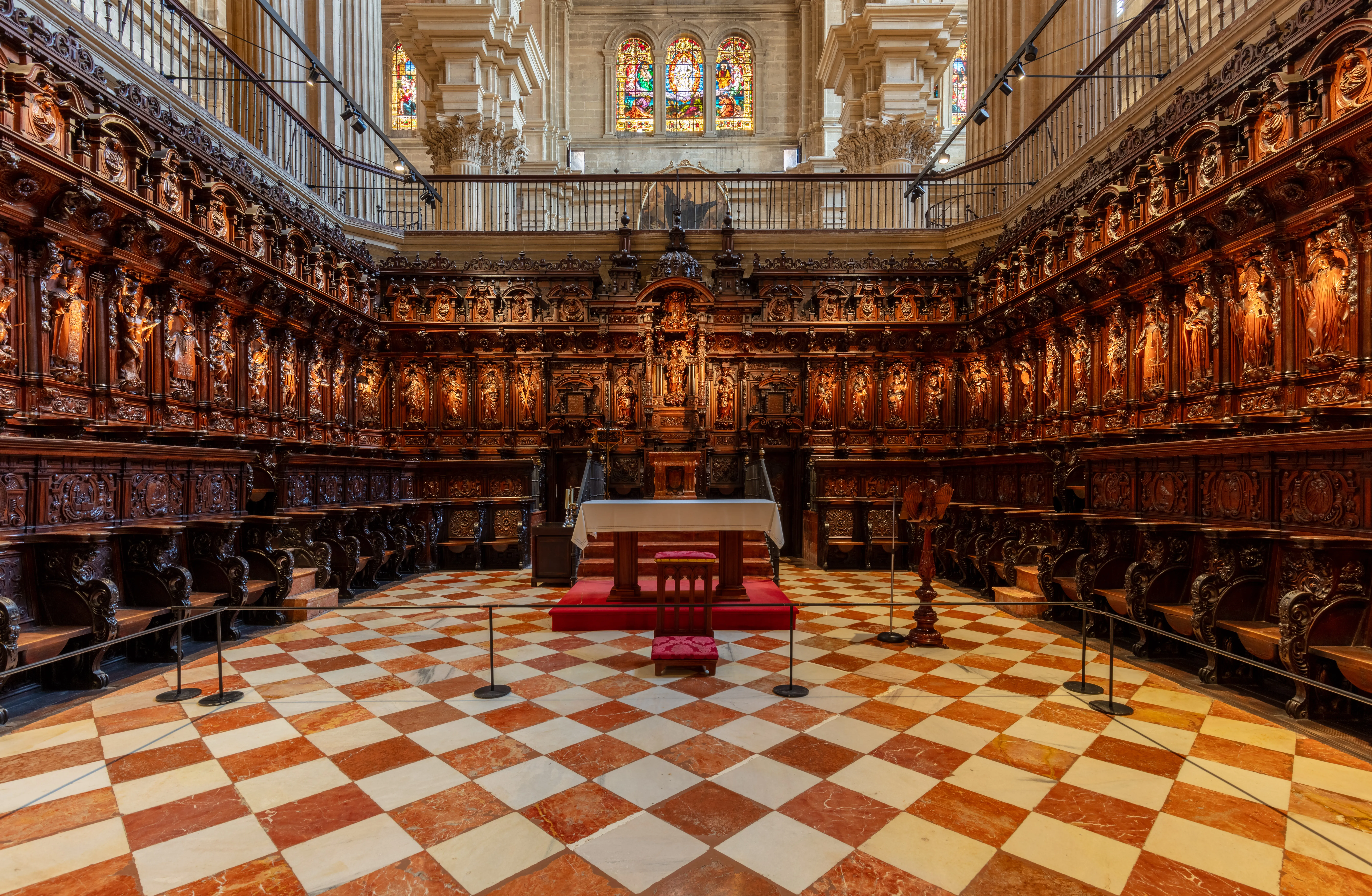 Sillerías de coro de madera ornamentadas con figuras talladas que bordean las paredes del interior de una catedral, rodeando un altar central sobre una plataforma roja con un suelo de mármol a cuadros.