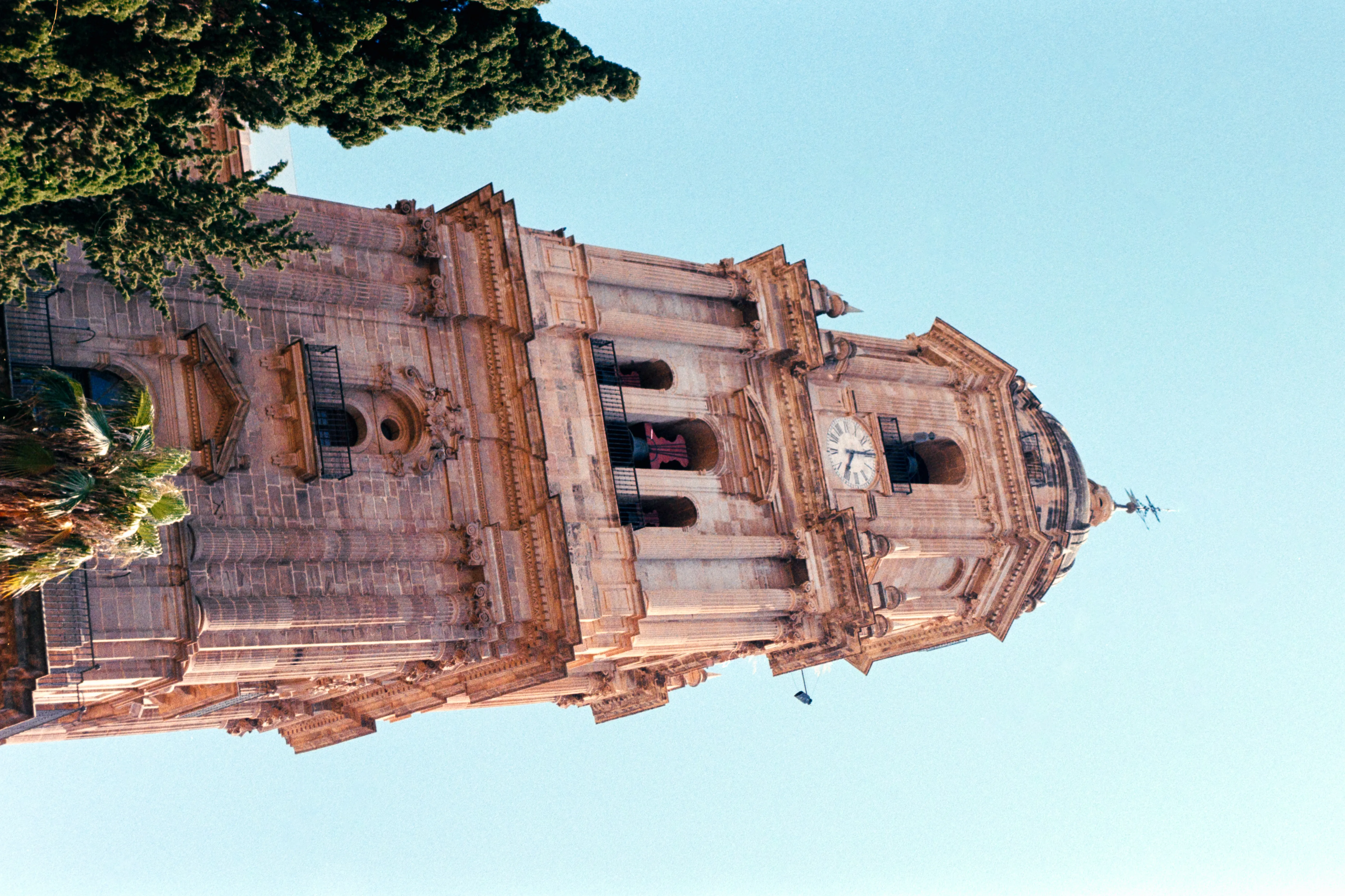 Torre de piedra barroca de la Catedral de Málaga con esfera de reloj, vanos arqueados para campanas y remate en cúpula sobre un cielo azul despejado, con un ciprés parcialmente visible