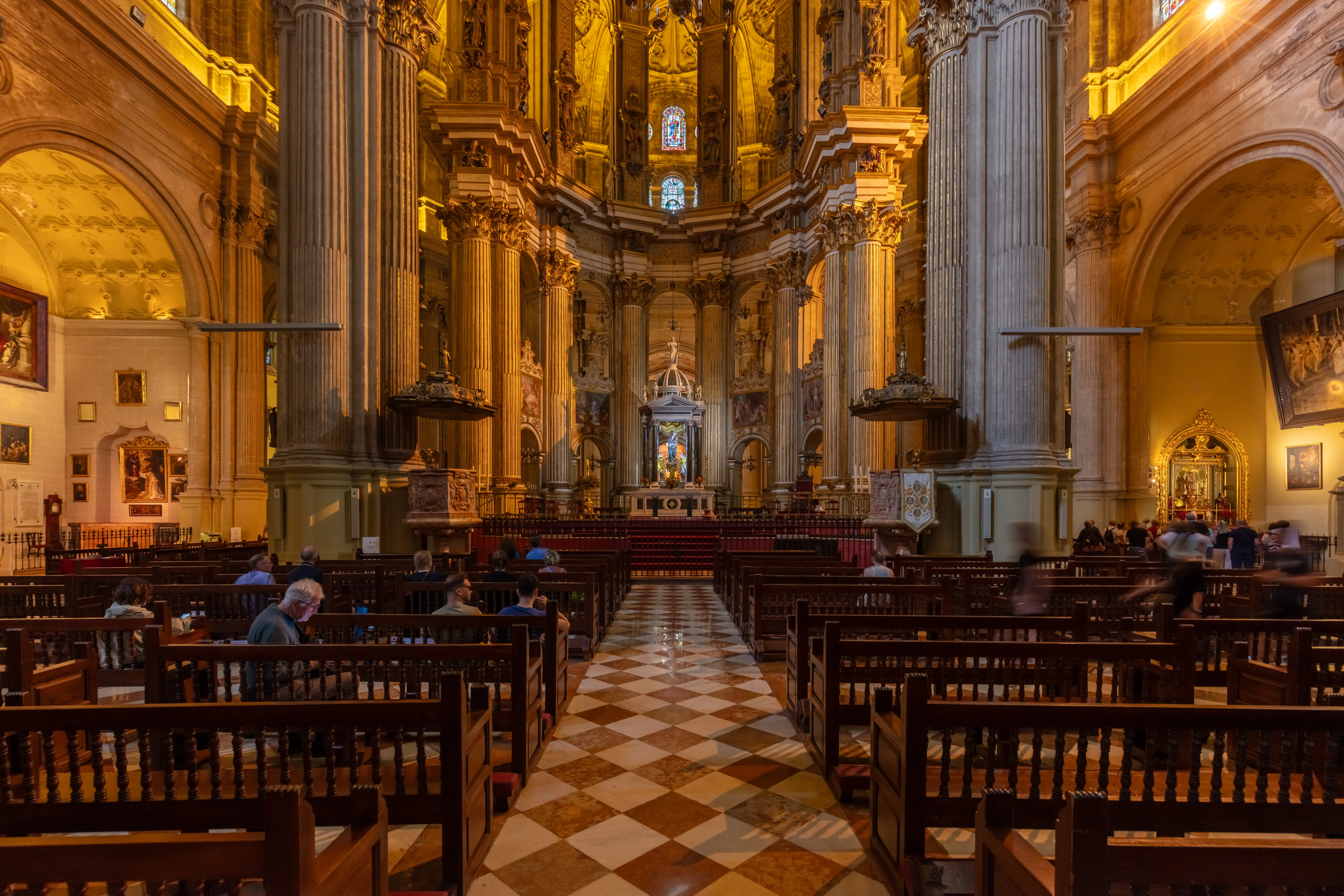 Vista interior de la Catedral de Málaga que muestra la nave con bancos de madera, suelo de mármol a cuadros, altas columnas de piedra y un ornamentado altar dorado al fondo.