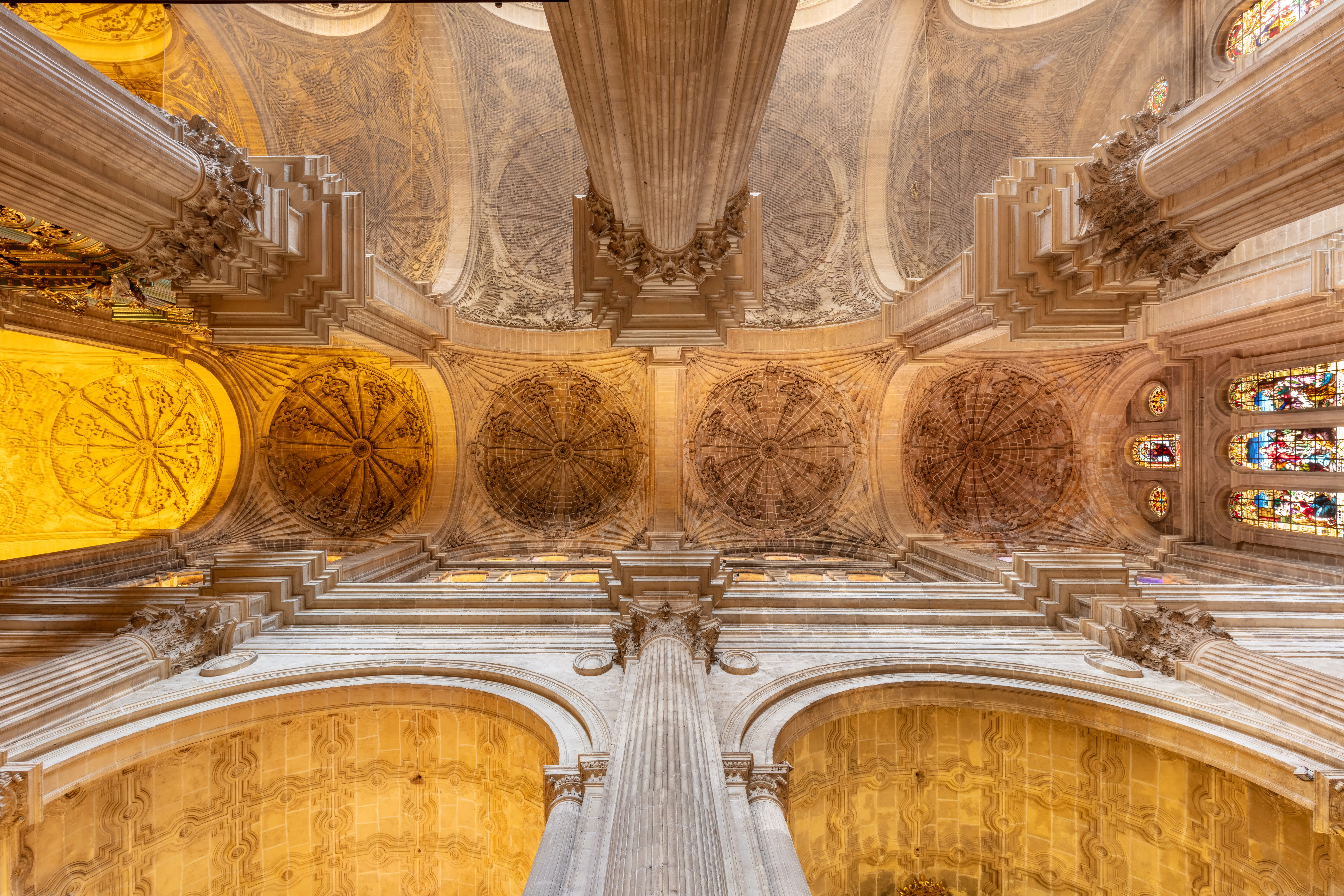 Techo abovedado de la Catedral de Málaga con decoraciones de piedra ornamentadas, medallones decorativos, columnas y vidrieras visibles desde abajo