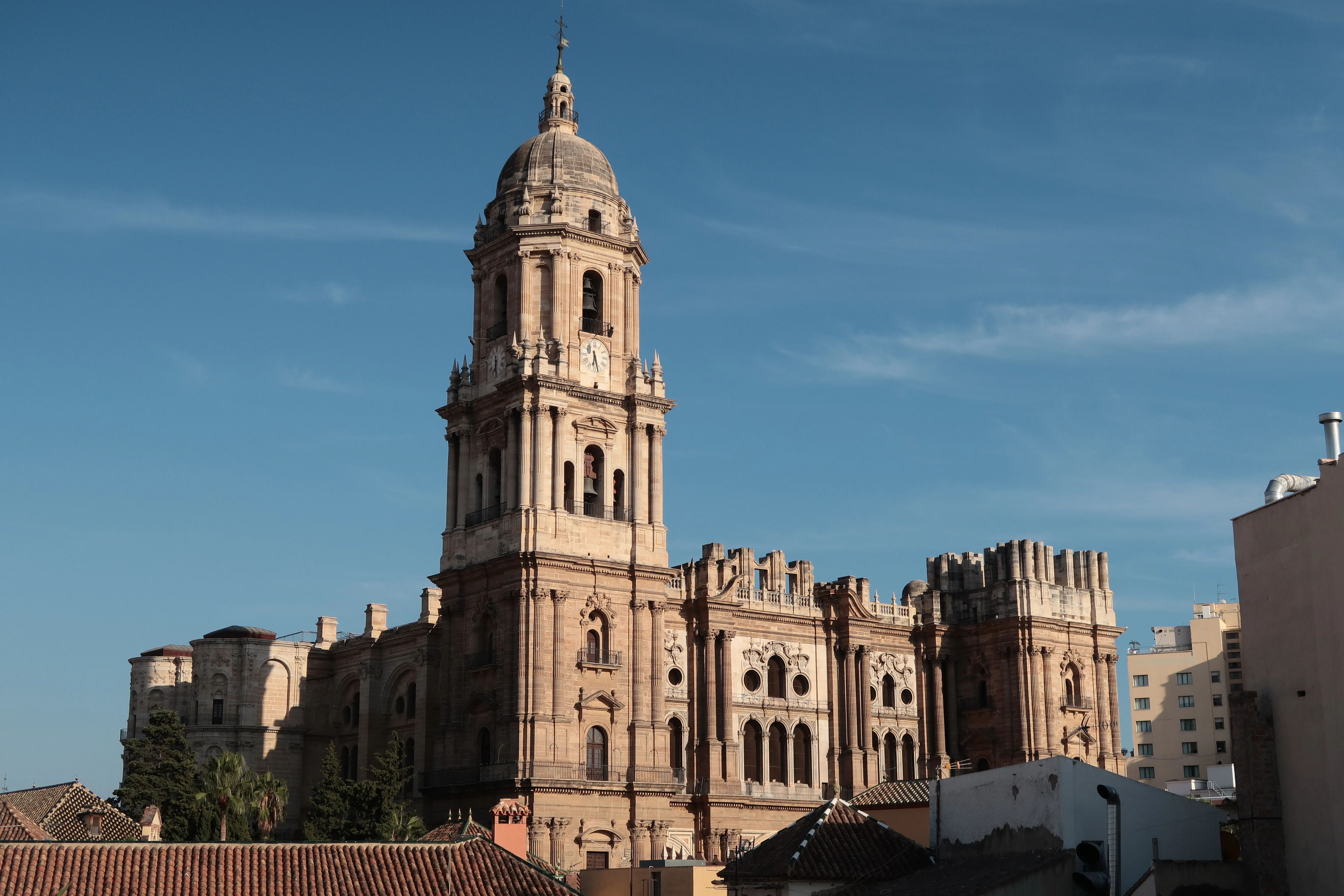 Vista exterior de la Catedral de Málaga mostrando su torre campanario terminada y la segunda torre inacabada contra un cielo azul, con tejados circundantes en primer plano