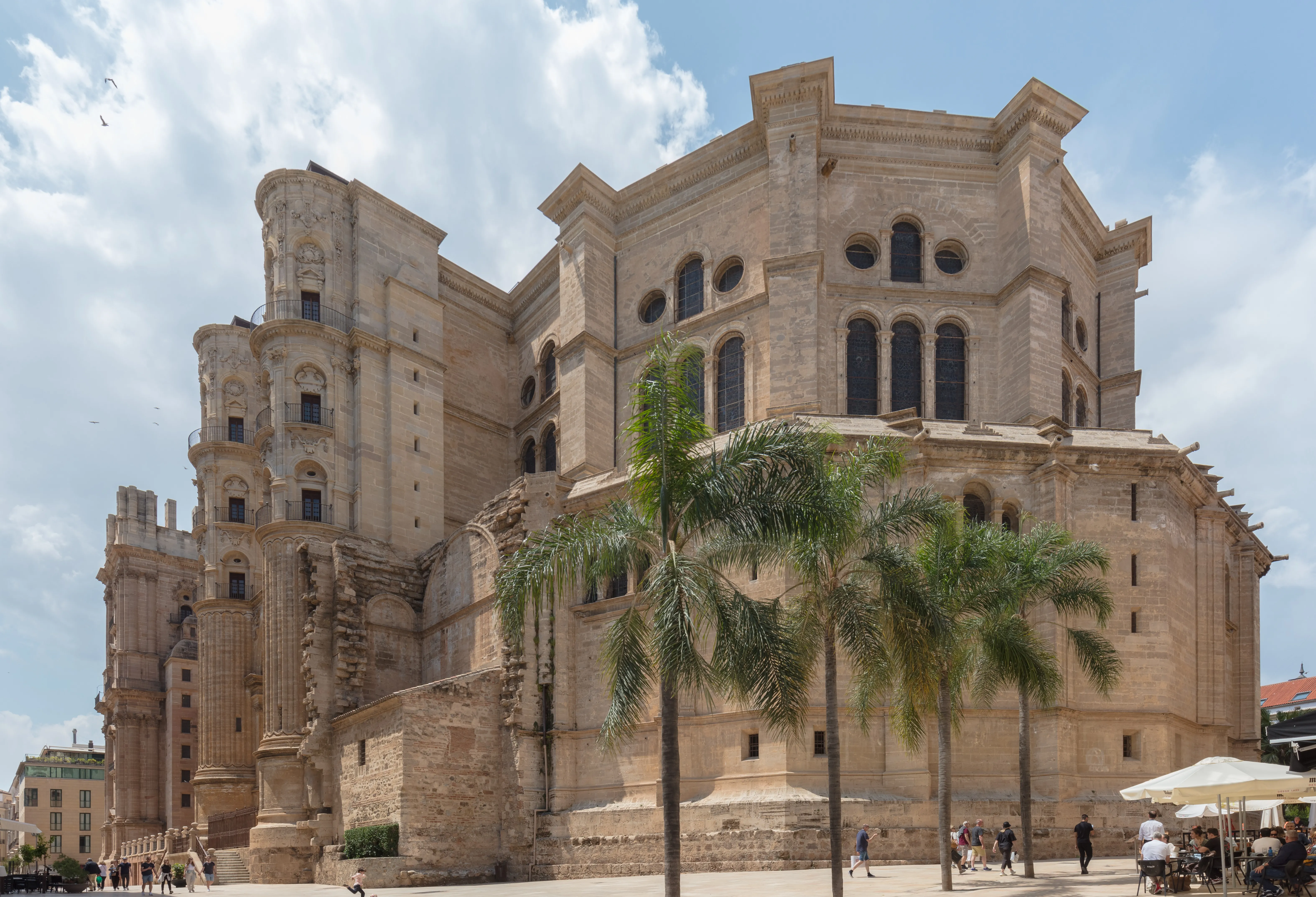 Vista exterior de la Catedral de Málaga mostrando el ábside de piedra redondeado, la torre inacabada y las palmeras en la plaza con visitantes paseando por debajo.