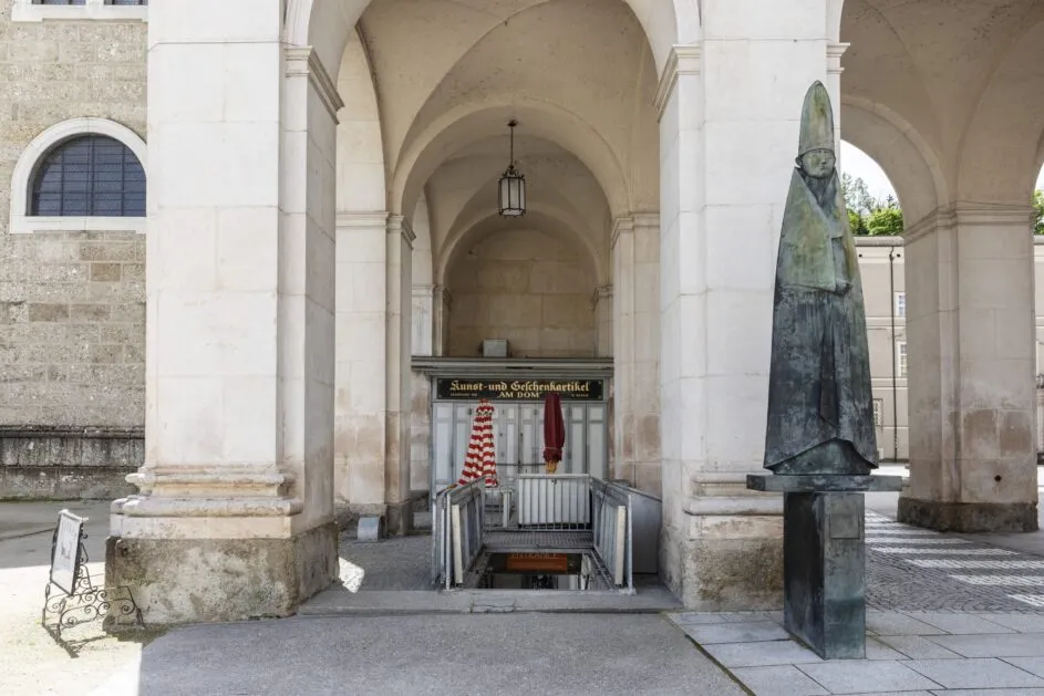 Columnata de arcos de piedra con una escalera de descenso que da entrada a un museo subterráneo, flanqueada por una estatua de bronce de un obispo a la derecha y un farol colgante arriba.