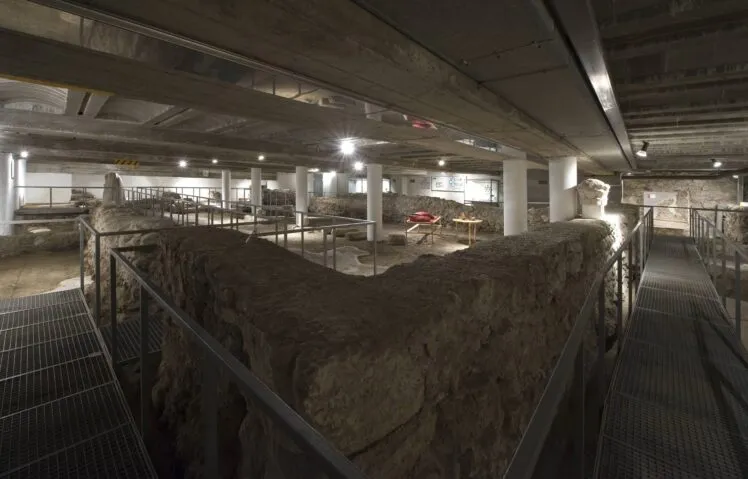 Sala de excavación arqueológica subterránea con antiguos muros de cimentación de piedra, techo moderno de hormigón con vigas y barandillas de metal en la pasarela del Cathedral Excavations Museum.