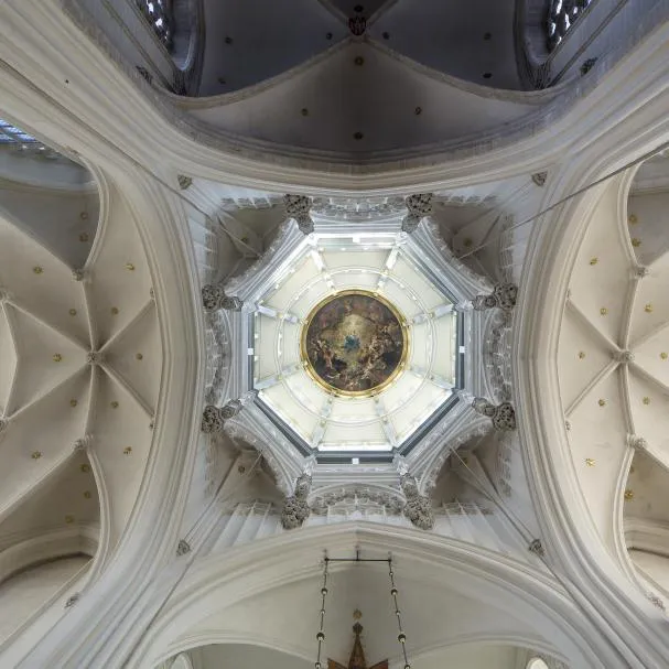 Vista hacia arriba de un techo abovedado octogonal blanco con una pintura circular central, rodeado de arcos abovedados en el interior de una catedral.
