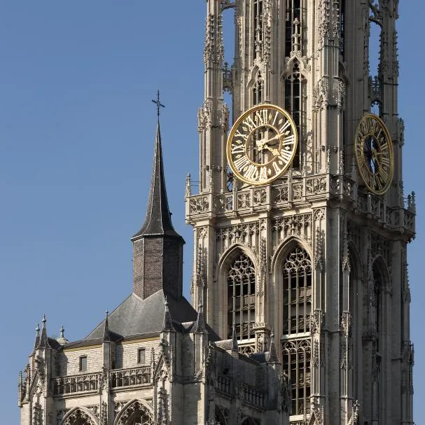 Torre de piedra gótica de la Catedral de Nuestra Señora en Amberes, con una gran esfera de reloj dorada y una aguja más pequeña y oscura contra un cielo azul despejado