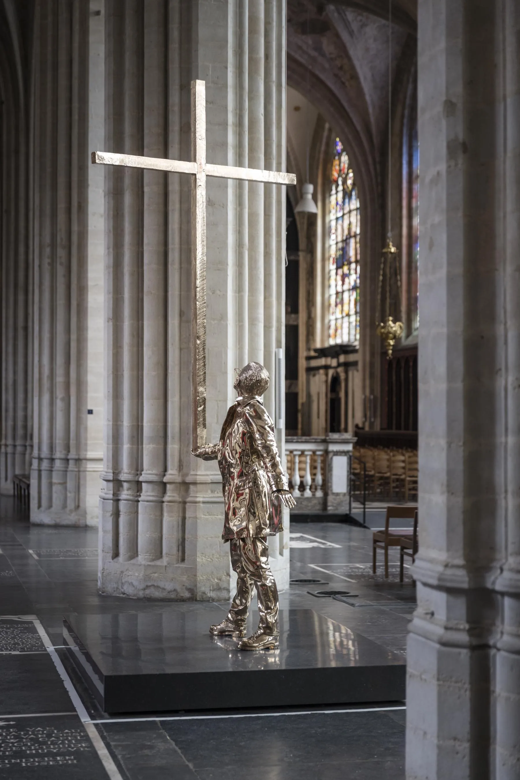 Una estatua dorada de un hombre cargando una gran cruz se encuentra sobre una plataforma negra dentro de una catedral gótica con altas columnas de piedra y vidrieras al fondo.