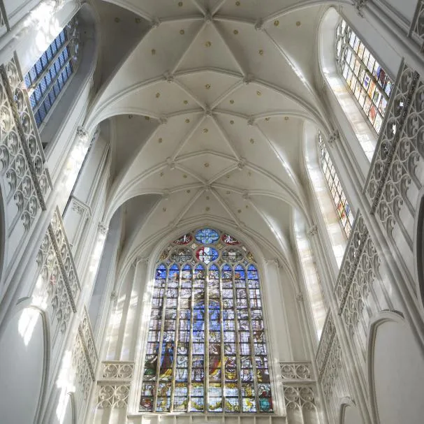 Vista en ángulo contrapicado hacia el techo abovedado de nervaduras blancas y un gran ventanal de vidrieras coloridas en el interior de una catedral gótica.