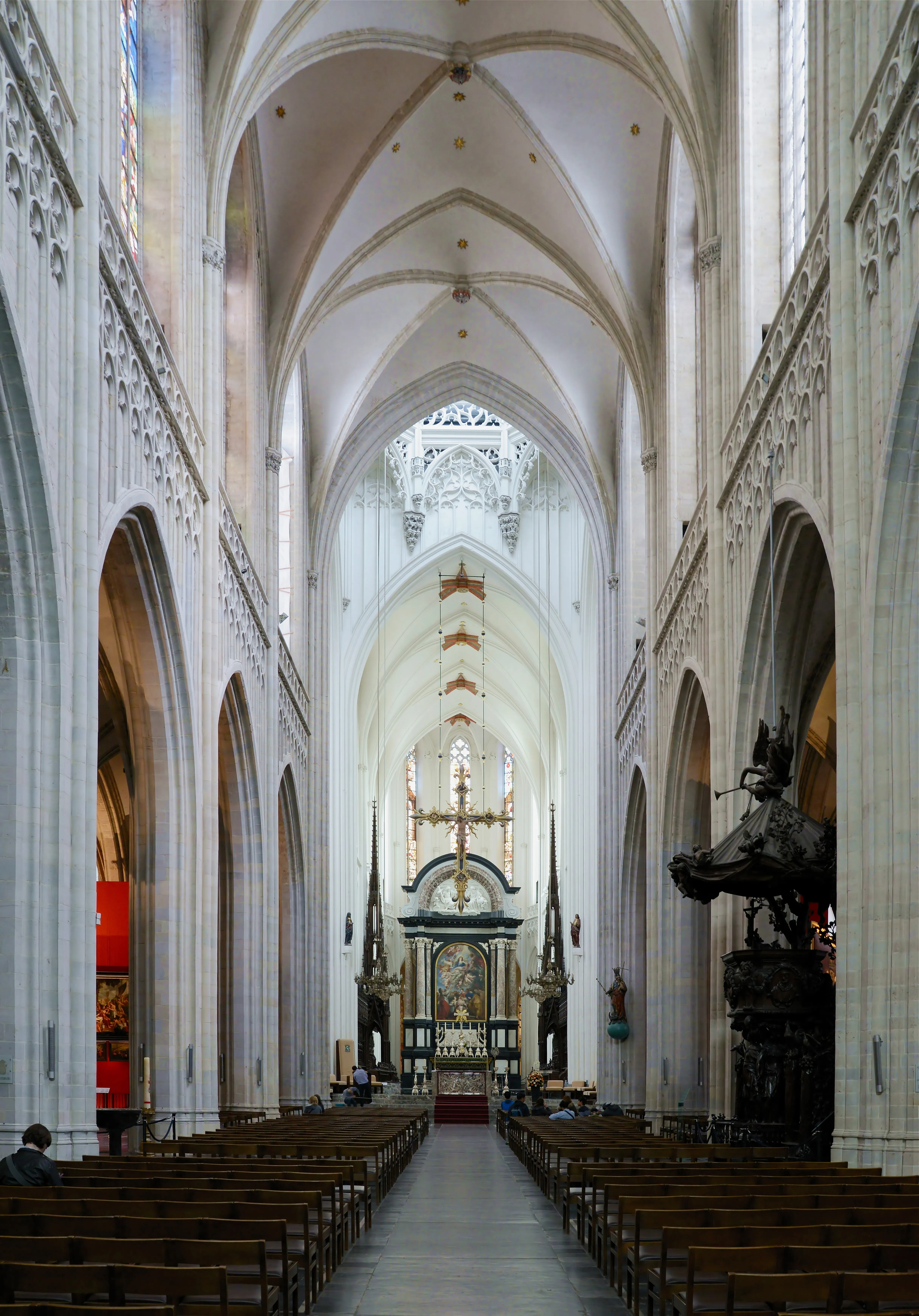 Nave larga de una catedral gótica con techos abovedados, hileras de bancos de madera y un altar con una gran cruz al fondo